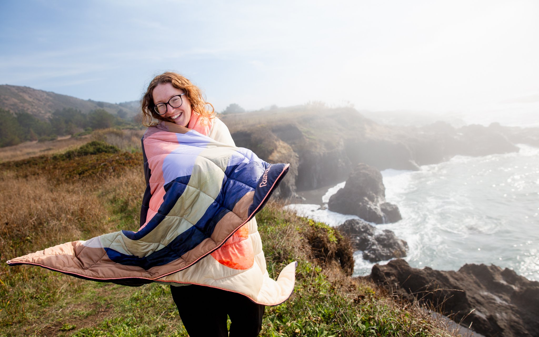 A woman with glasses smiling, holding a colorful quilt, standing near a coastal cliff with ocean waves crashing below and a foggy sky in the background.