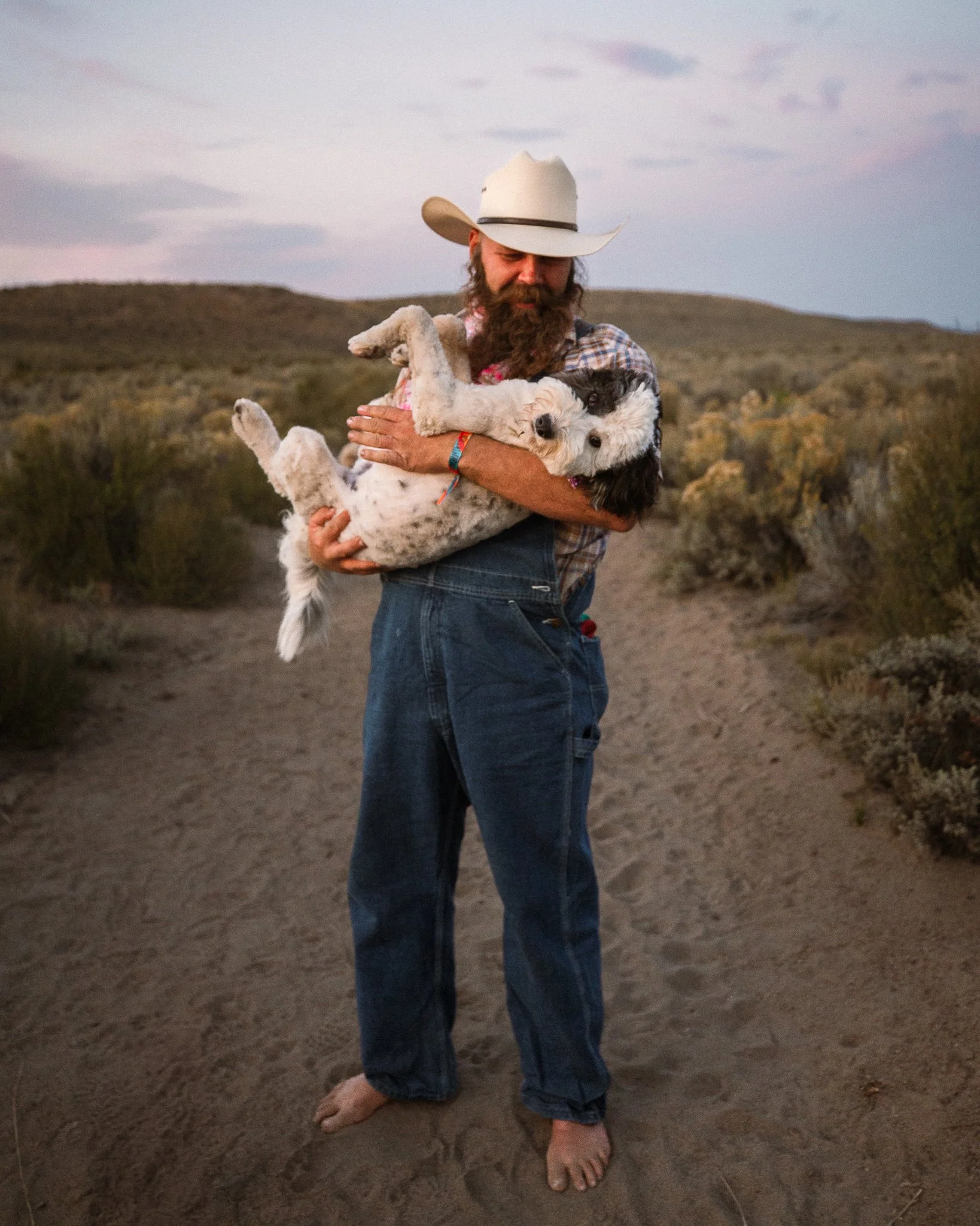 A man with a cowboy hat, beard, and jeans stands barefoot on a dirt path in a desert landscape, holding a small white dog with black markings in his arms.