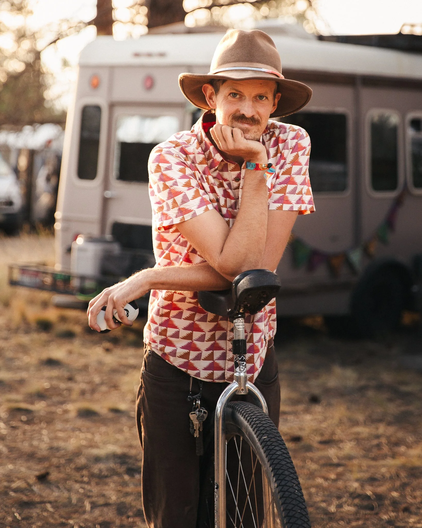 A man with a mustache wearing a brown wide-brimmed hat and a pink patterned short-sleeve shirt, resting his chin on his hand while leaning on a unicycle, in front of a vintage camper trailer with a string of colorful flags, outdoors during sunset.