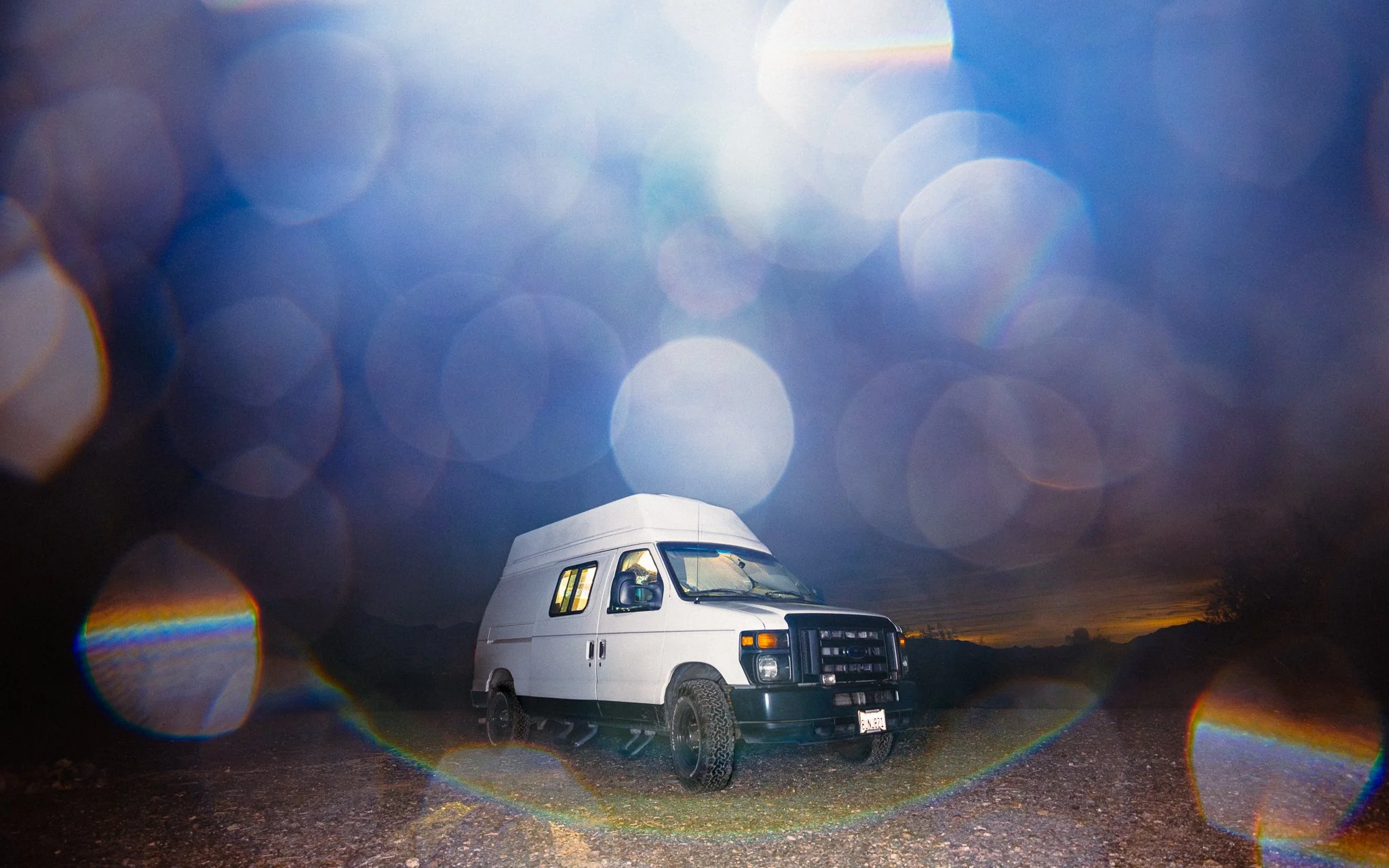 A white van parked on a dark, rocky terrain at dusk or night with a colorful bokeh effect and lens flare in the foreground.