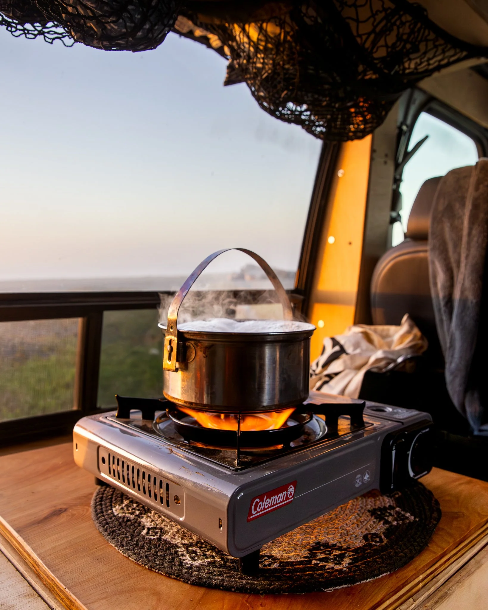 A steaming pot on a portable Coleman stove inside a camper van, with a landscape view through the window during sunset or sunrise.