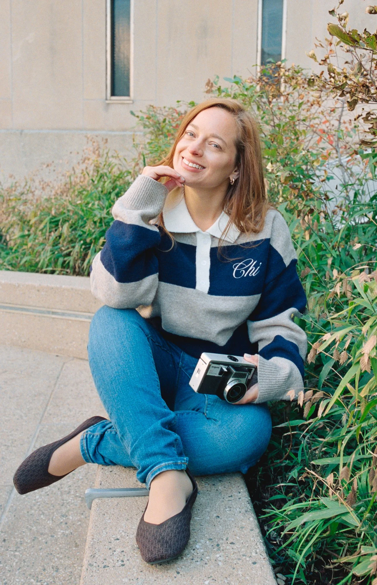 A young woman with red hair and fair skin sitting on a concrete bench outdoors, holding a vintage camera, smiling, wearing a navy and gray striped sweater with 'Chi' embroidered on it, blue jeans, and black flats, with lush green and brown foliage behind her.