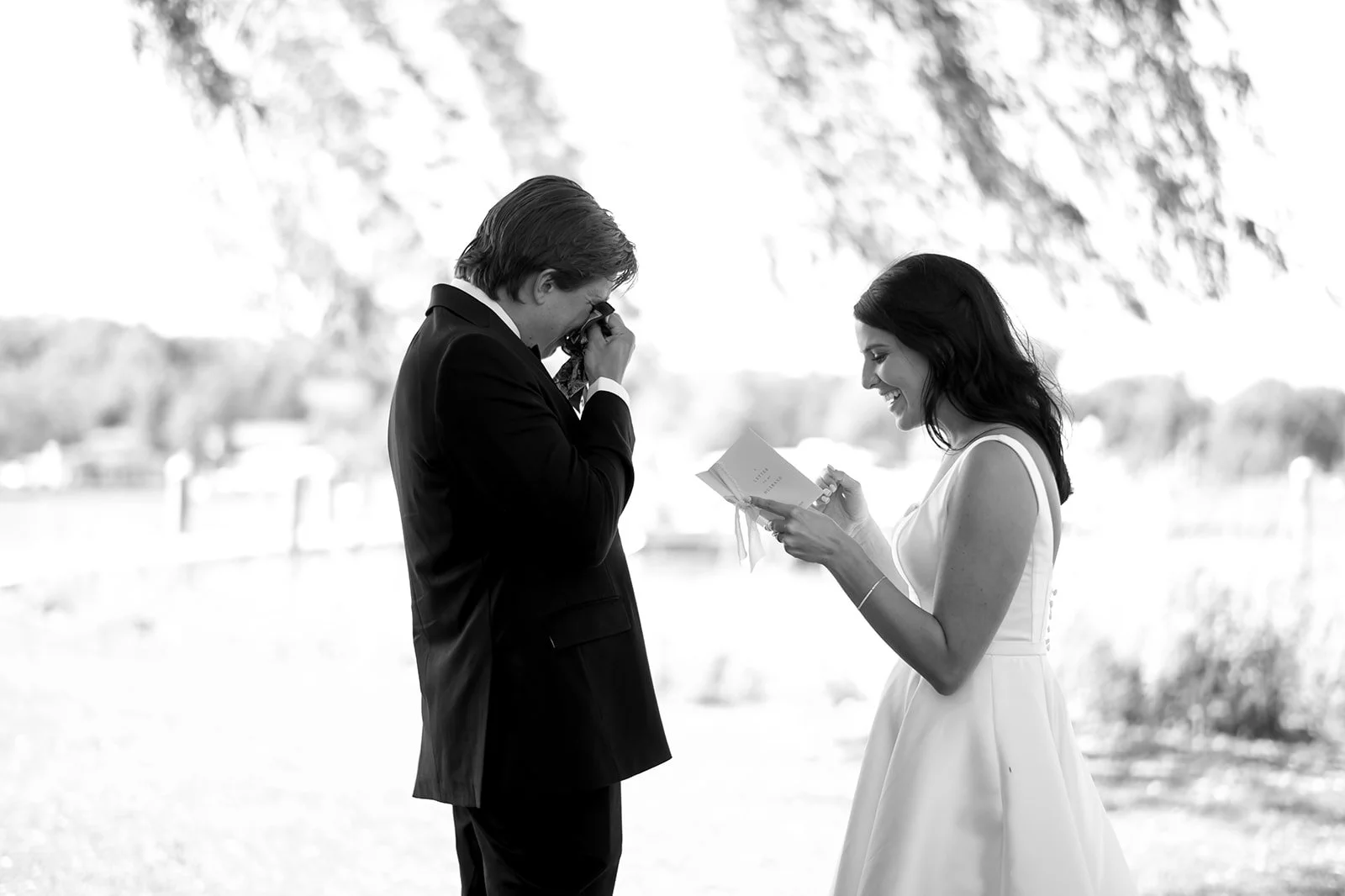 A black and white photo of a wedding ceremony outdoors showing a man in a suit wiping his eyes, possibly crying, while a woman in a wedding dress reads from a paper, smiling.