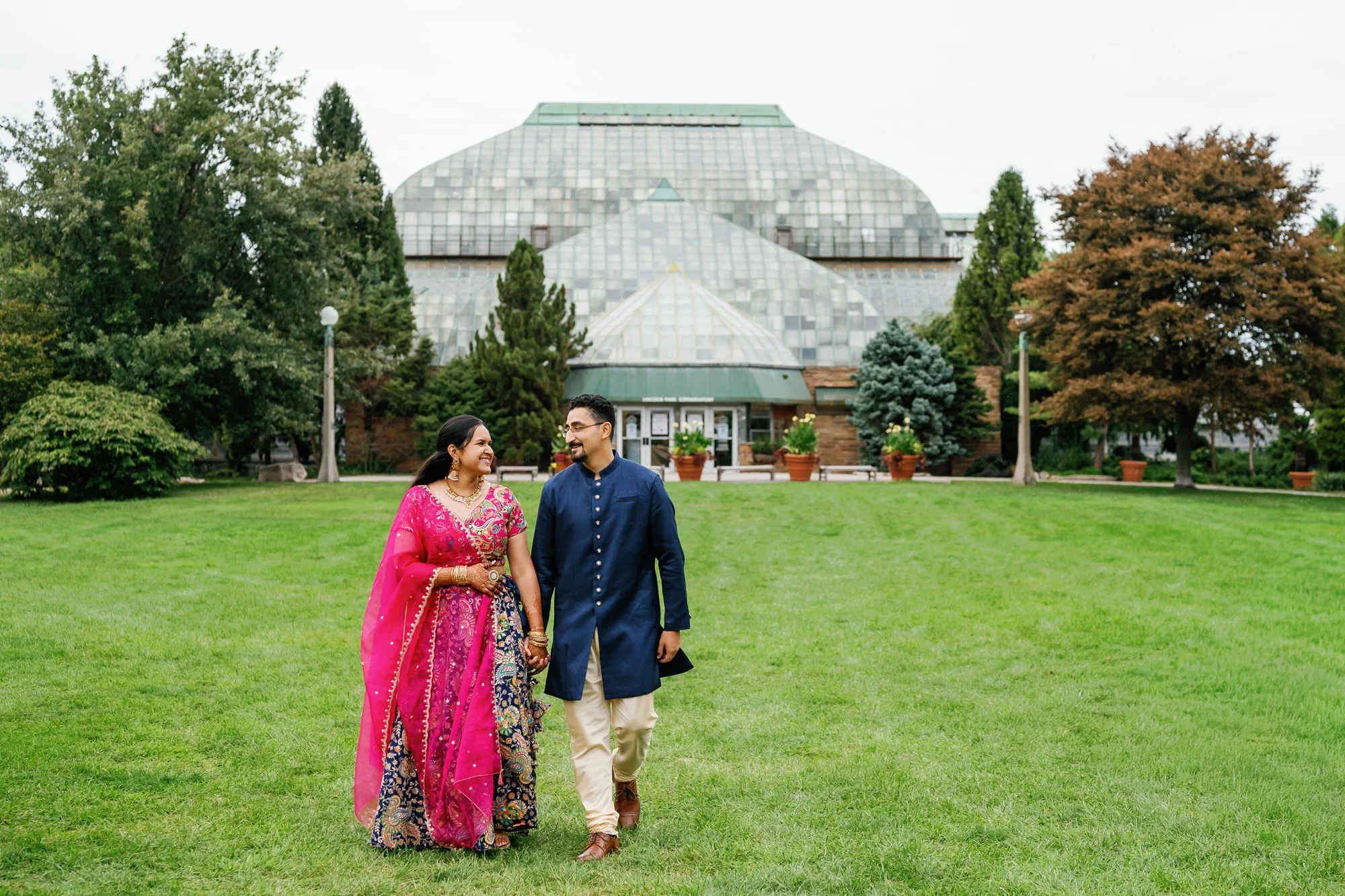 Couple in traditional South Asian clothing posing on a lawn in front of a large glass conservatory