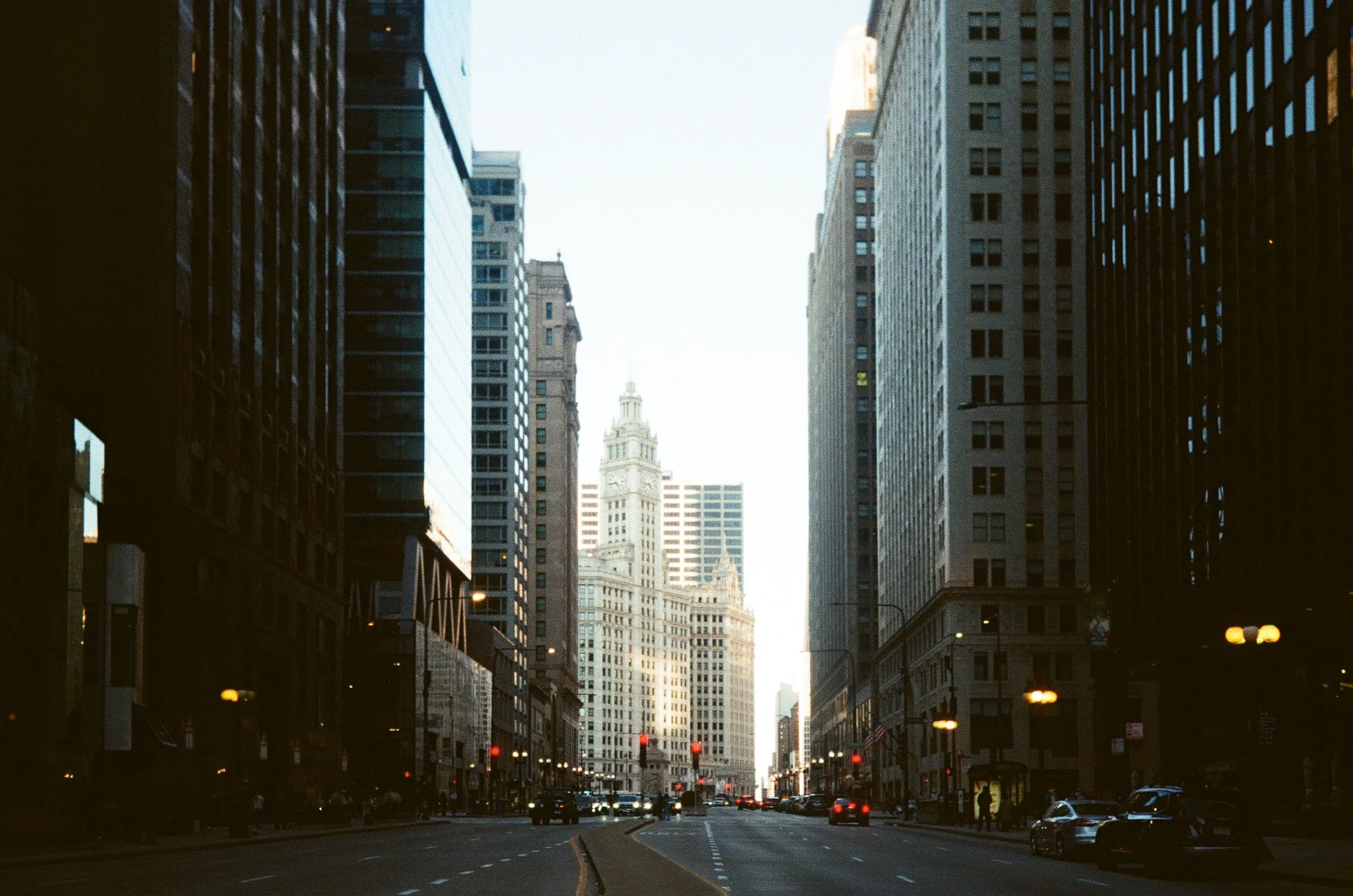 A city street in downtown with tall skyscrapers on both sides, cars driving, and a clock tower building in the distance.