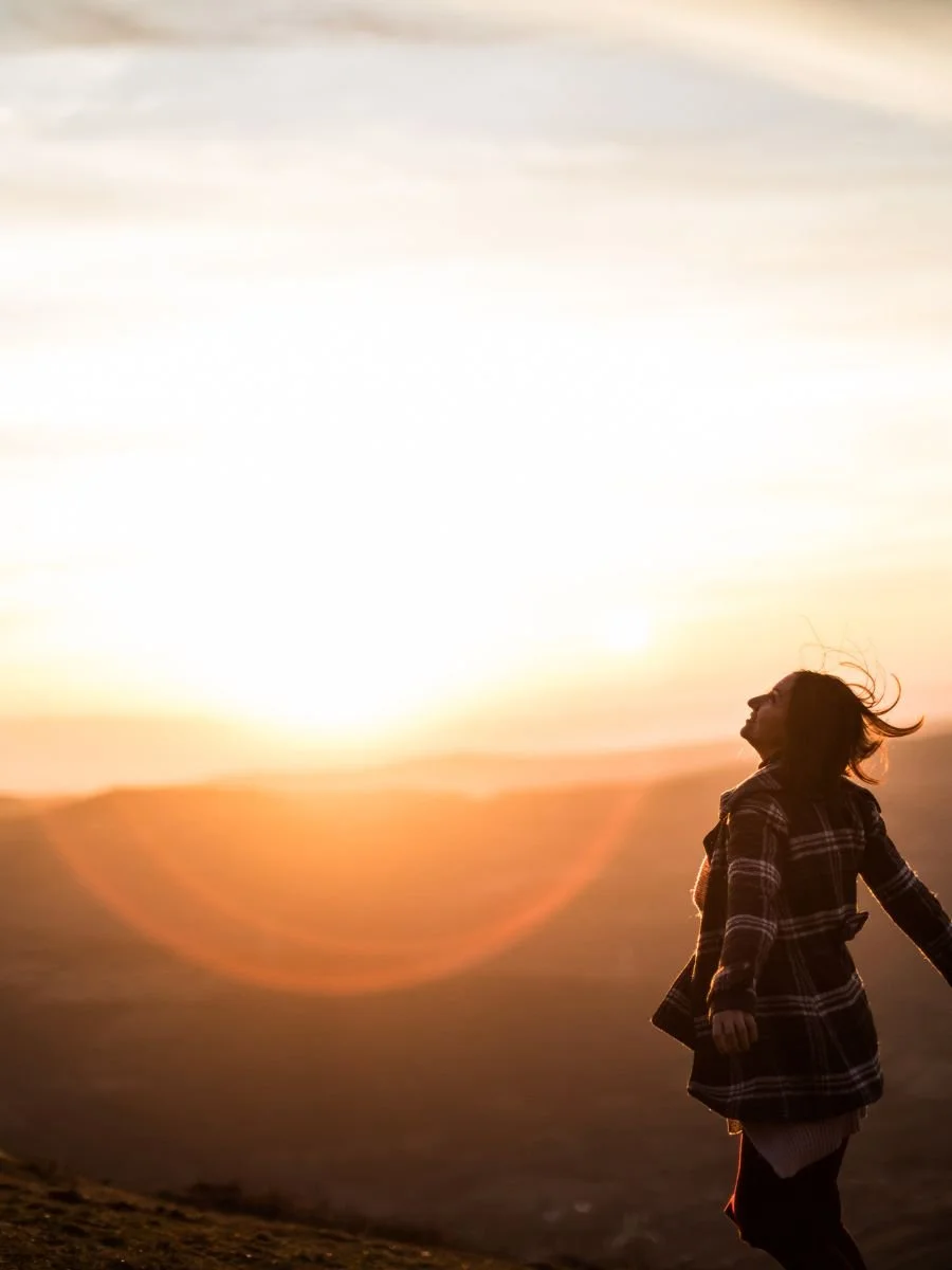 A person standing outdoors during sunset with arms outstretched, wearing a plaid jacket and enjoying the scenery.