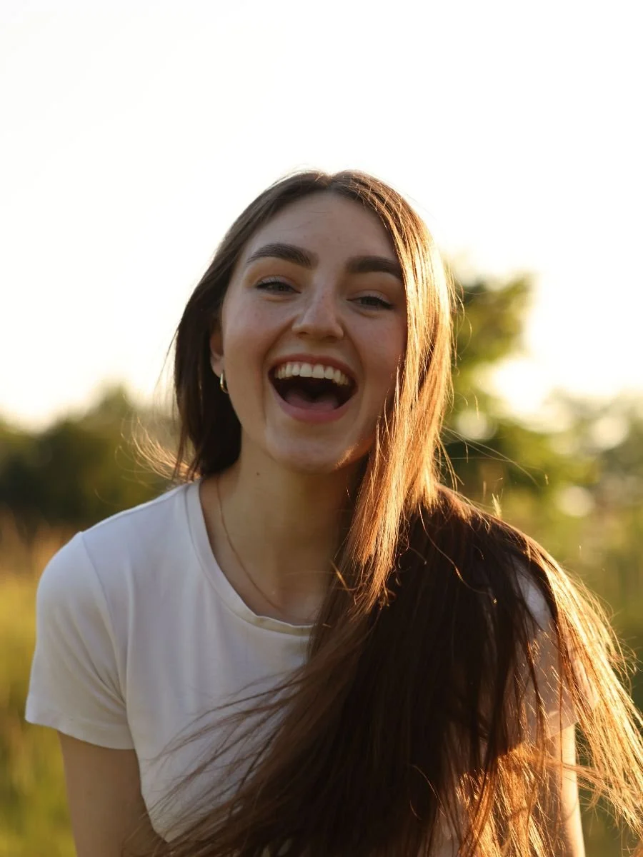 Young woman with long brown hair, smiling and laughing outdoors during sunset, wearing a white t-shirt.