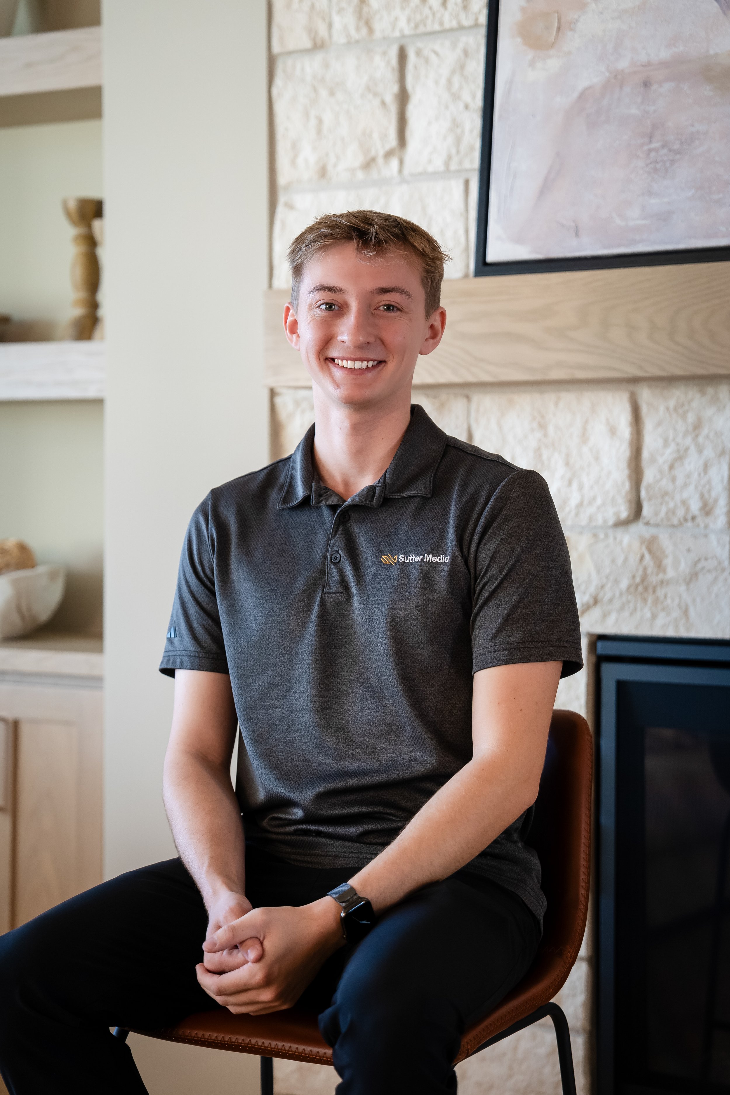 Young man with short light brown hair wearing a navy blue t-shirt, smiling, against a plain light grey background.