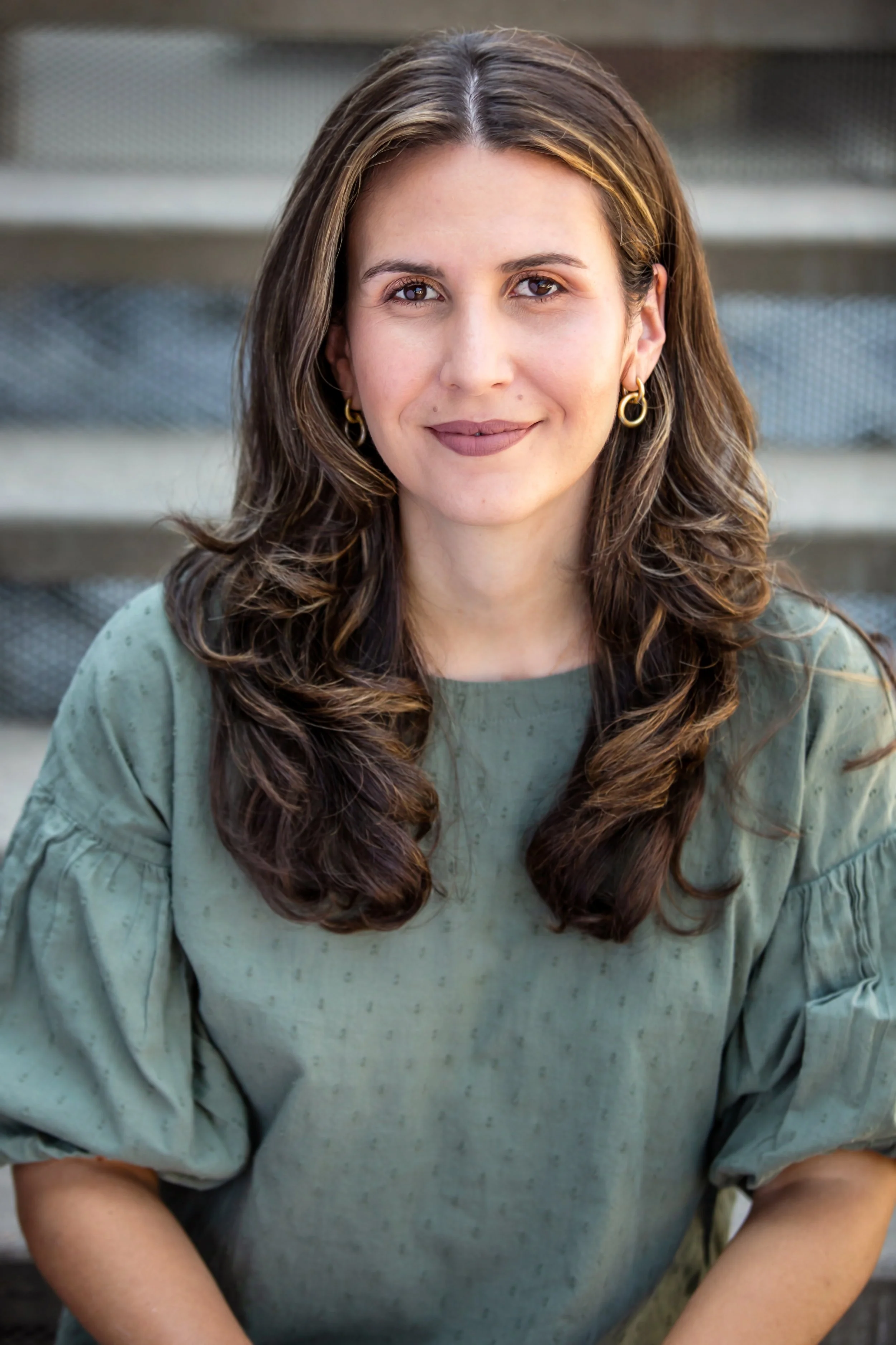 A woman with long, wavy brown hair and hoop earrings, wearing a green top, sitting outdoors with blurred bleacher seats in the background.