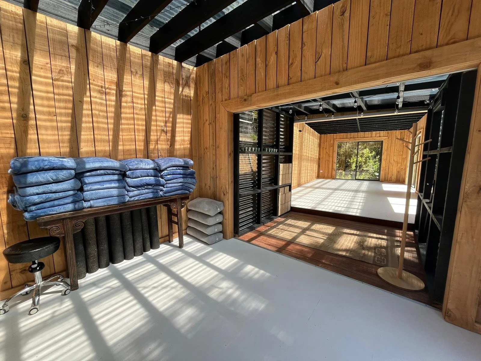 Interior of a wooden room with folding beds, storage shelves, and a sliding glass door leading outside, with sunlight creating shadows on the floor.