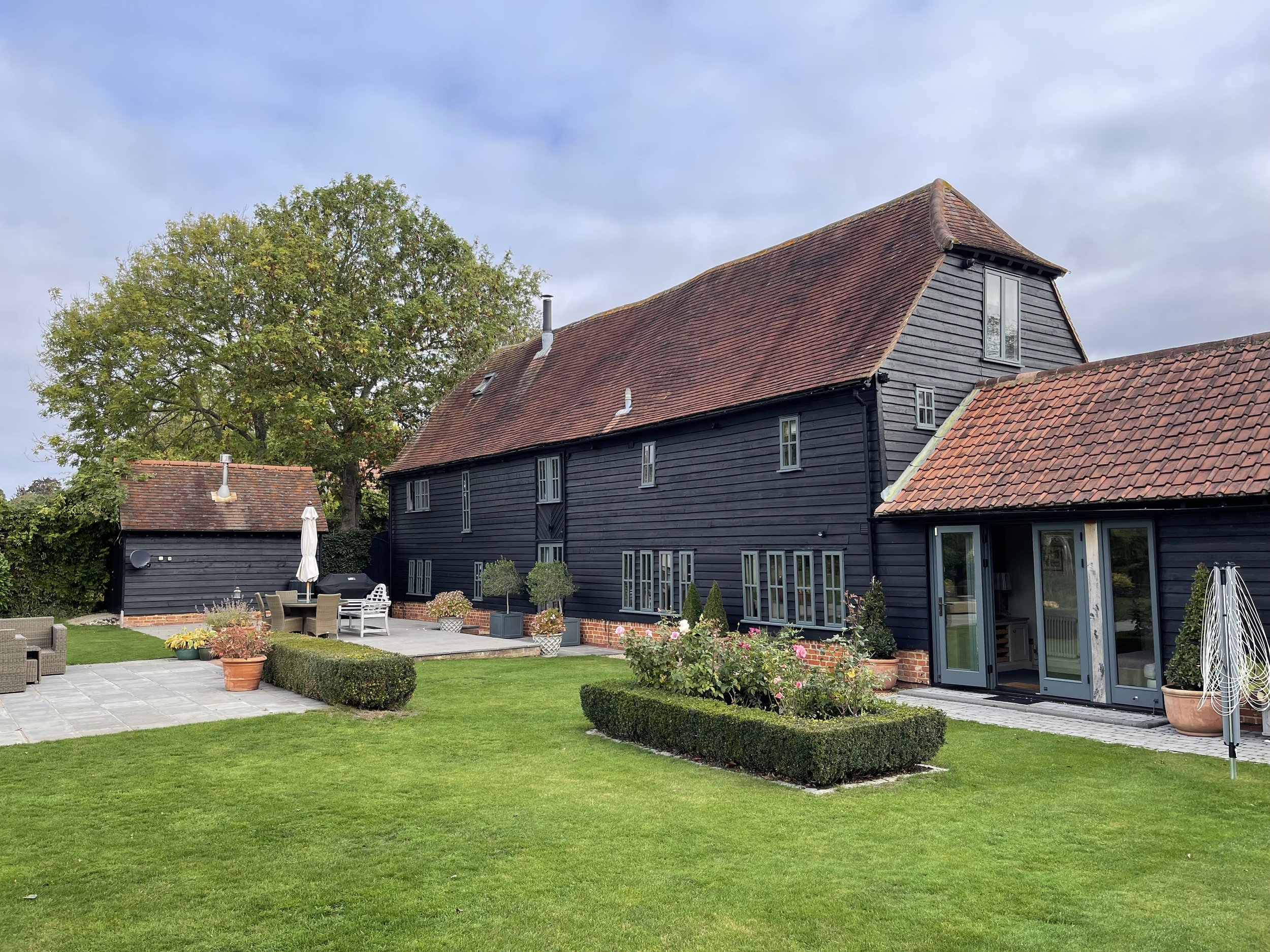 A large black wooden barn-style house with red tile roof, gray doors, and small windows in a well-maintained garden with green lawn, potted plants, hedge, patio area, outdoor chairs, and umbrella, with a large tree and cloudy sky in the background.