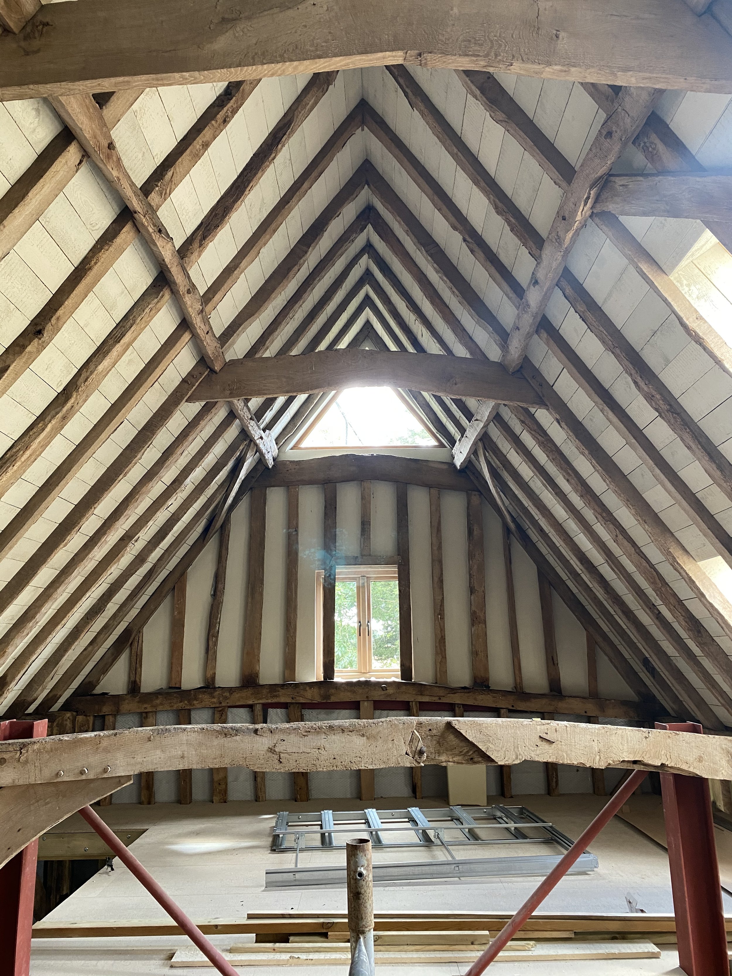 View of an unfinished attic with exposed wooden beams and insulation, featuring a small window and a skylight at the peak of the ceiling.