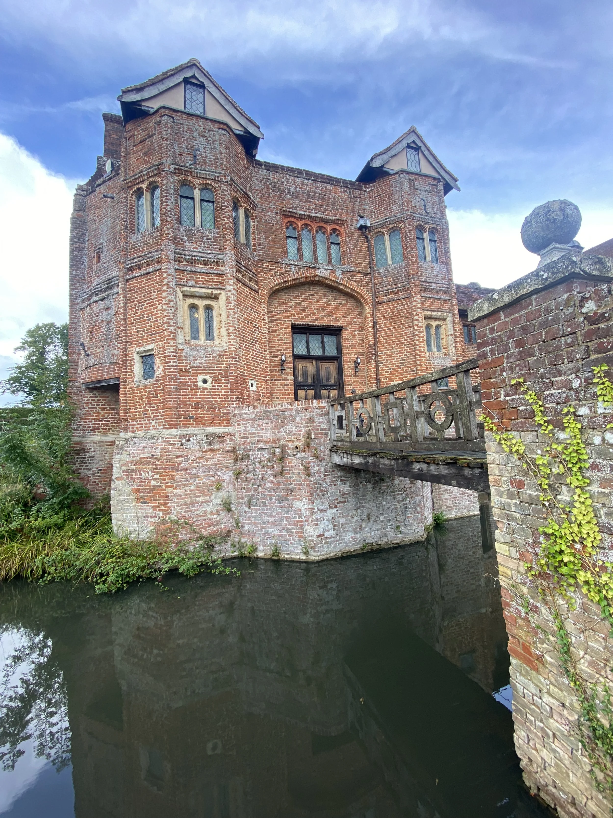 A historic brick castle with pointed towers and small windows, situated beside a water canal with a small bridge and brick wall, under a partly cloudy sky.