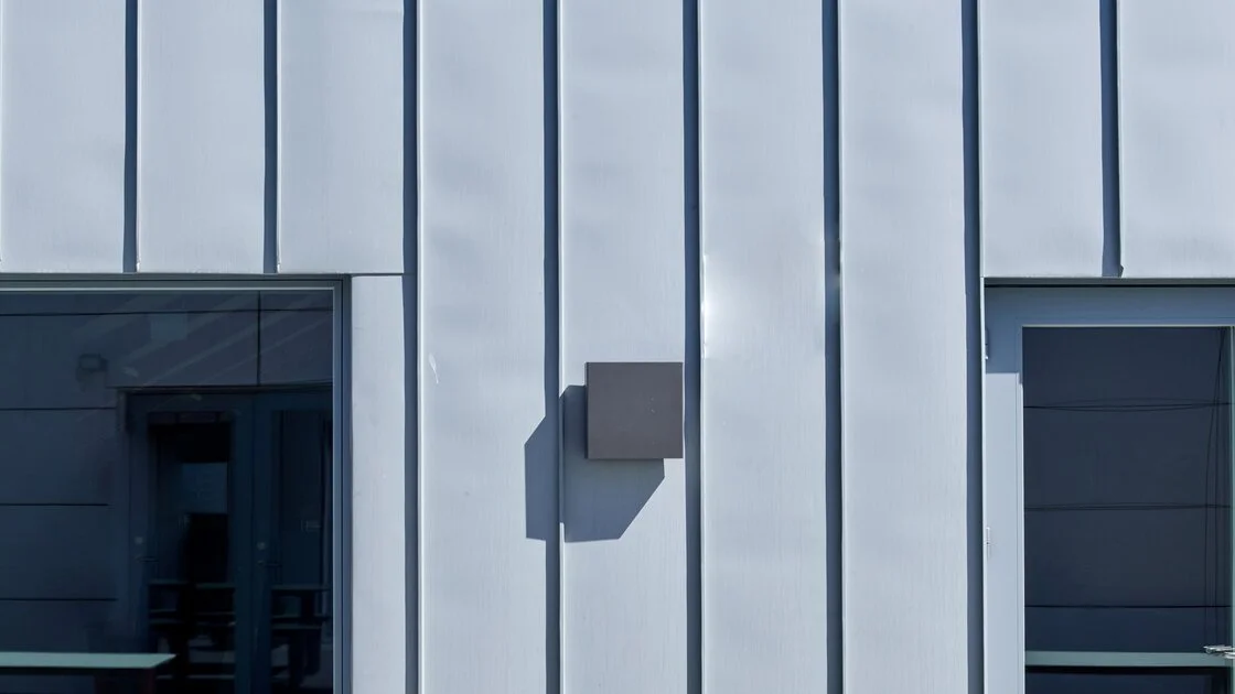 Close-up of a modern building's exterior with vertical metal panels, two windows, and a box-shaped external fixture casting a shadow.