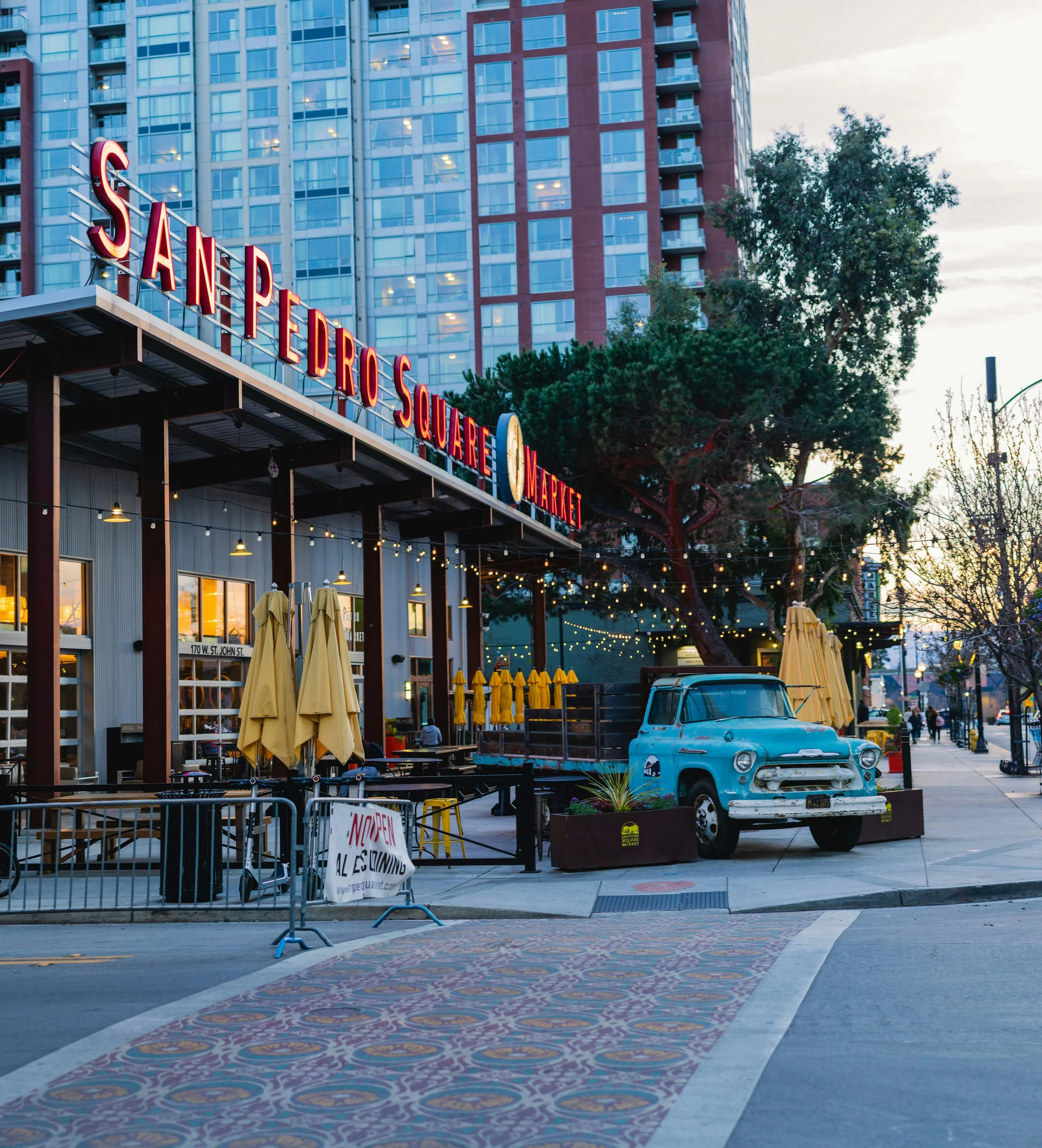 Outdoor scene of the Sanchez Square Market with yellow umbrellas closed and stacked outside, a vintage blue truck parked on the street, and a high-rise building with many windows in the background during early evening.