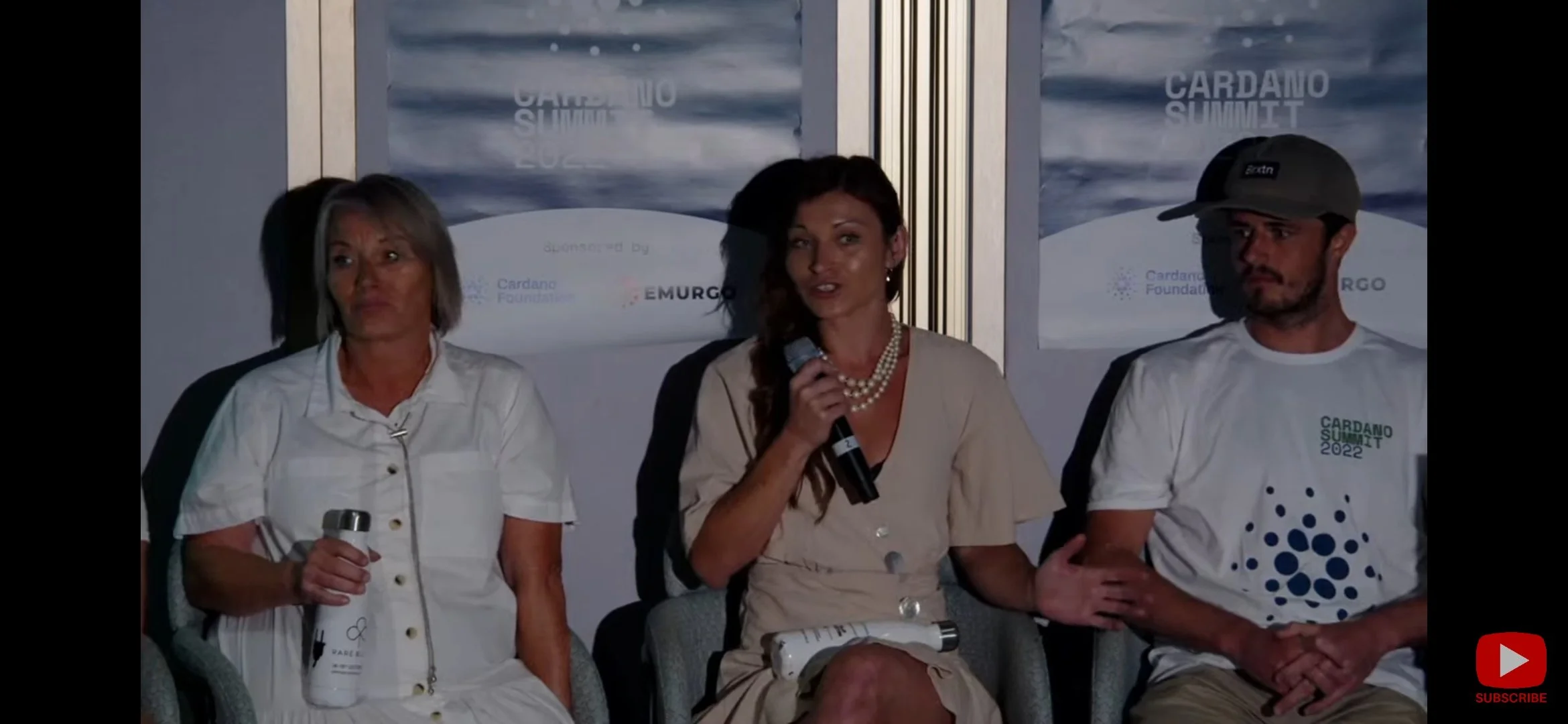 Three people seated at a panel discussion at the Cardano Summit 2022. One holds a microphone while speaking. Visible event logos and banners in the background.