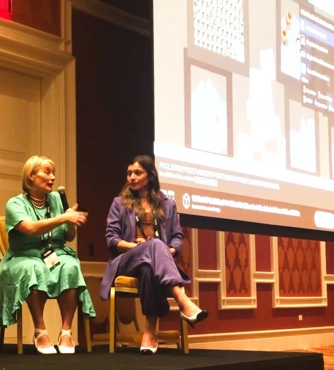 Two women sitting on stage, engaging in a panel discussion at a conference, with a large screen displaying content in the background.