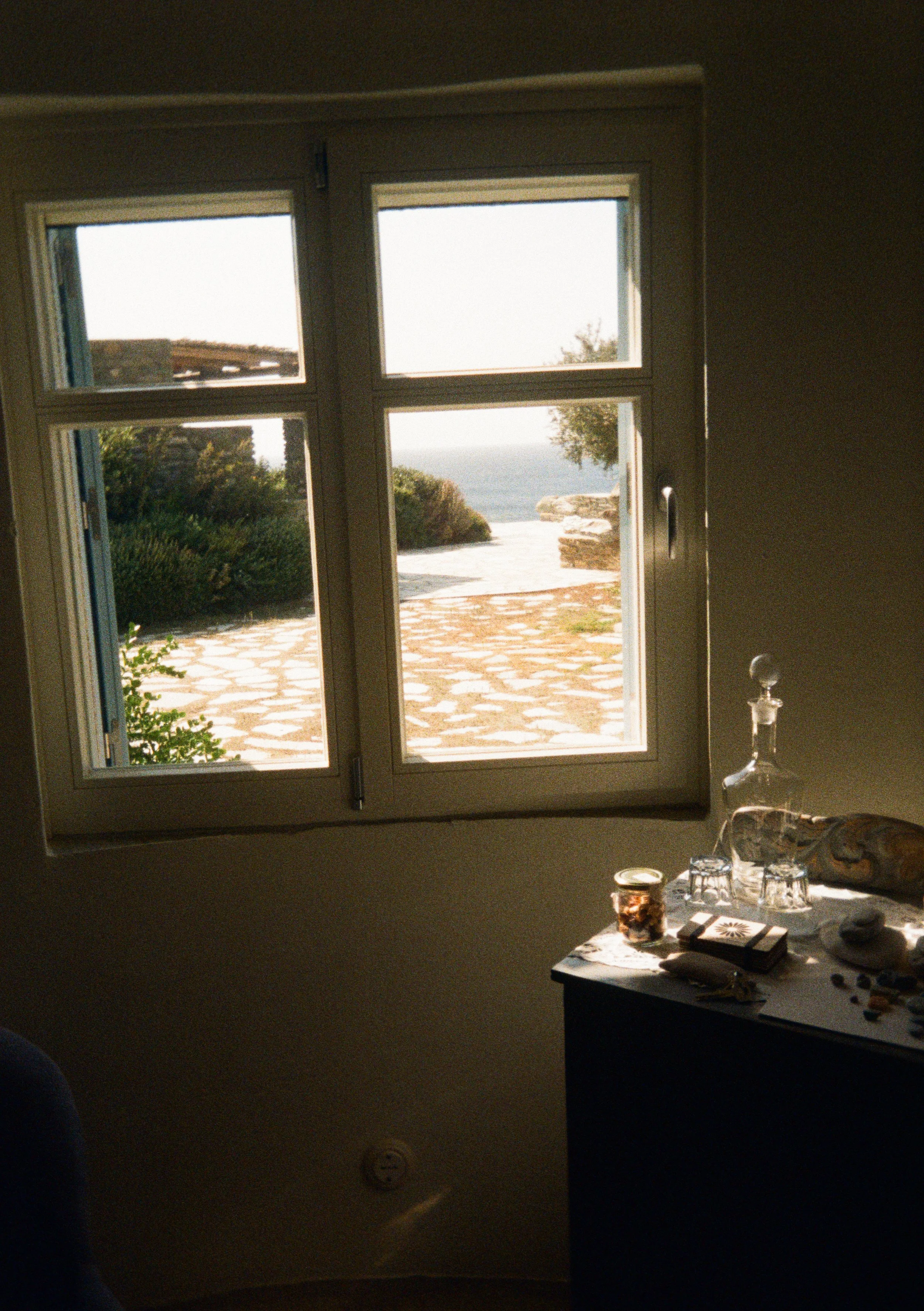 View through a window showing a stone-paved outdoor area, bushes, and a body of water in the distance.