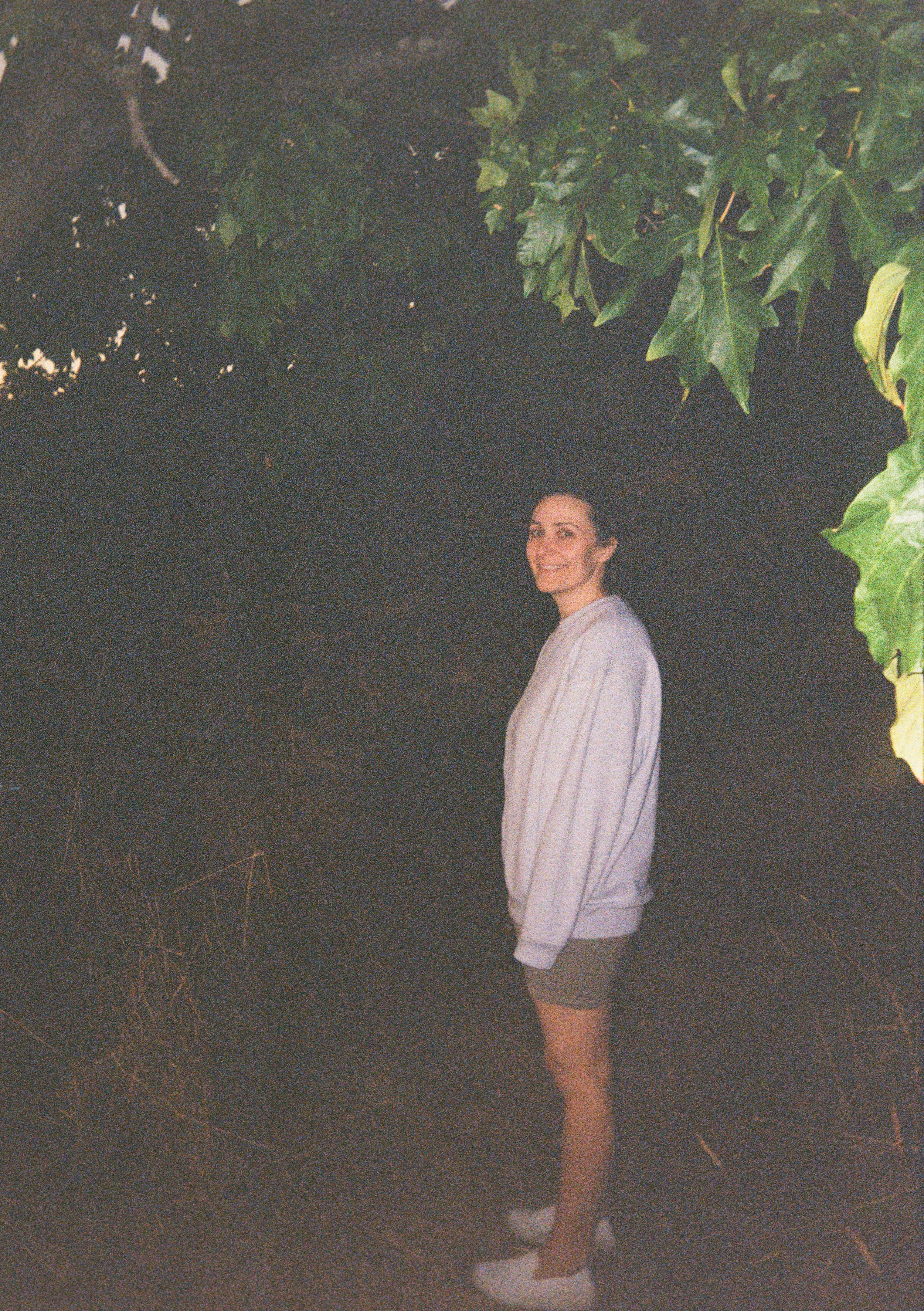 A woman standing outdoors at night, smiling, with dark foliage above and behind her, dressed in a light-colored sweater, shorts, and white shoes.