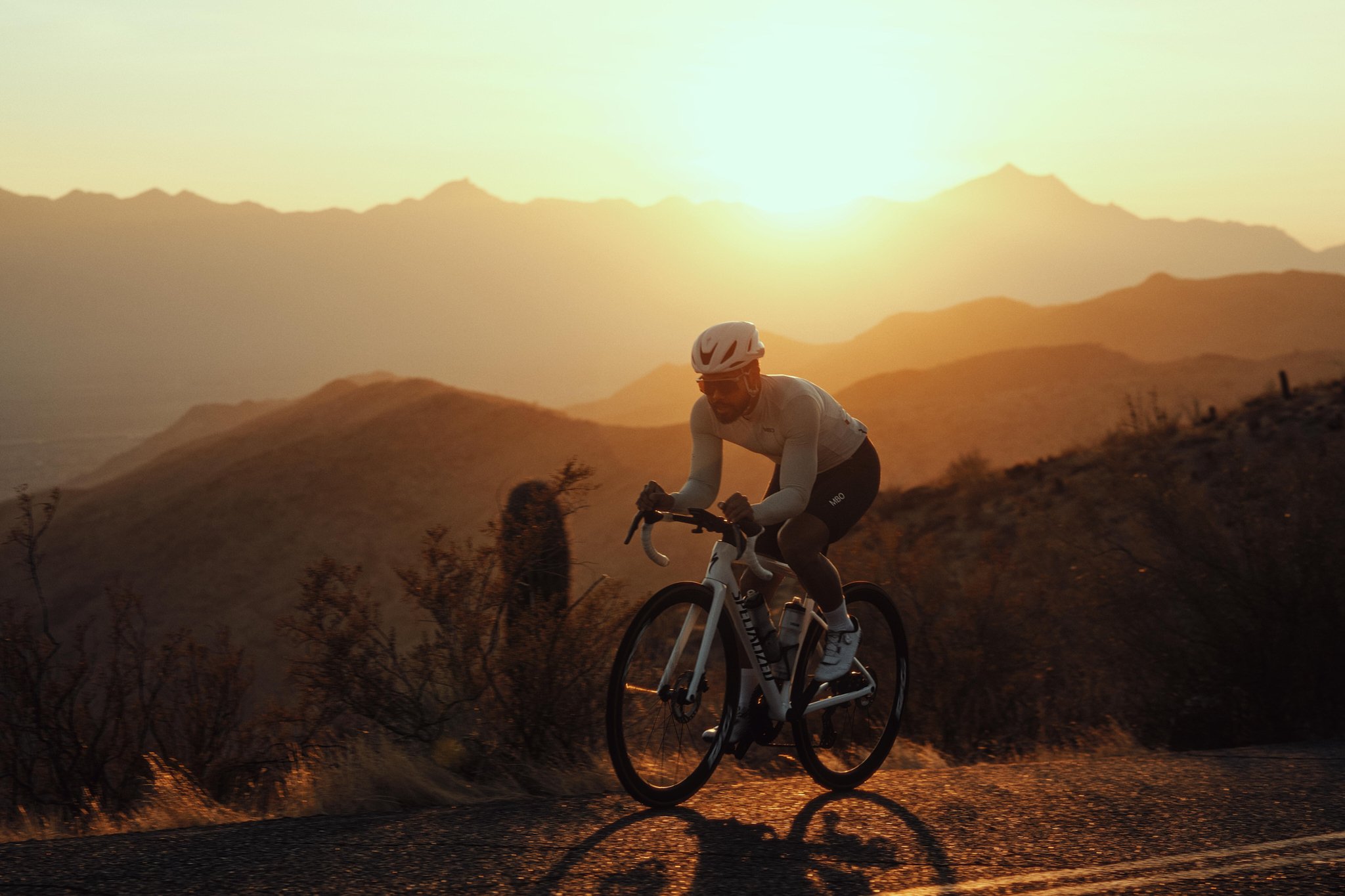 A man riding a bicycle on a mountainous road during sunset with a scenic backdrop of hills and mountains.