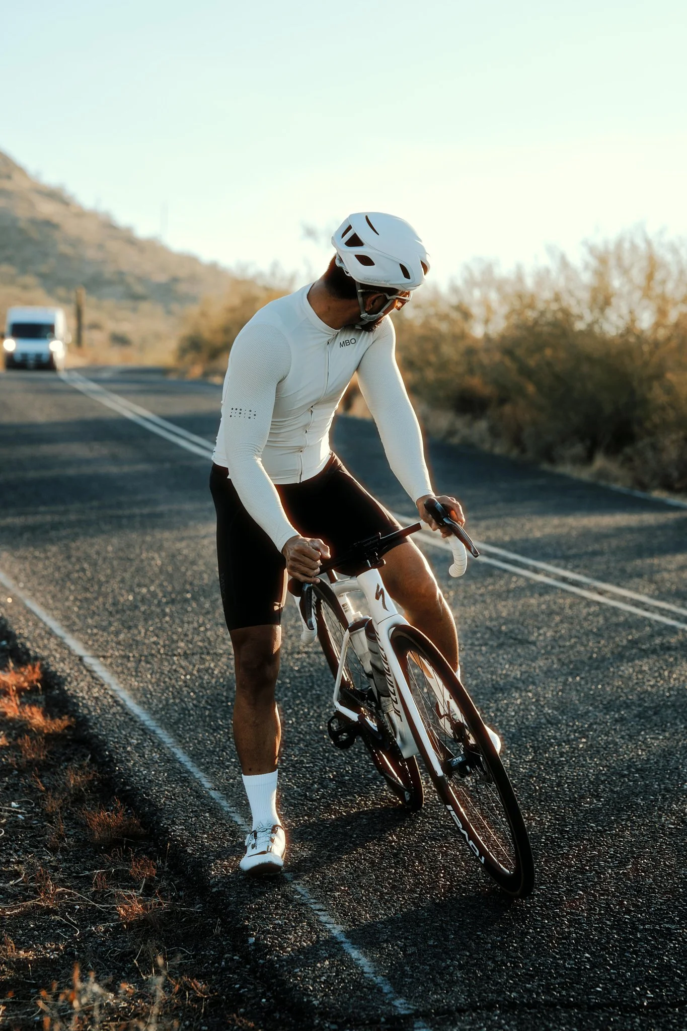 A man in a white helmet and athletic clothing riding a bicycle on a paved road in a desert landscape during sunset.