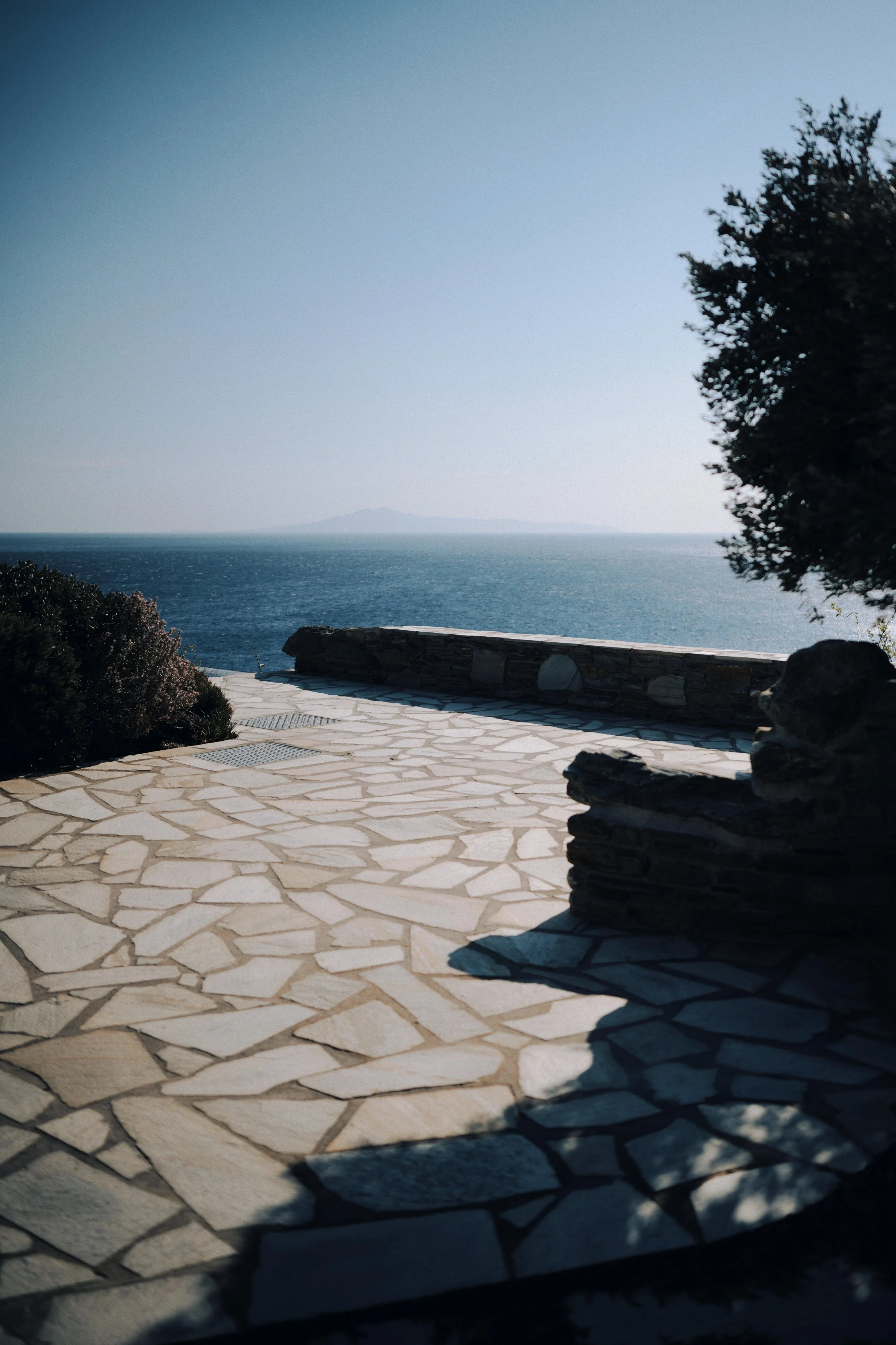 A stone patio with a low stone wall overlooking the ocean, with trees and bushes on each side and an island in the distance under a clear blue sky.