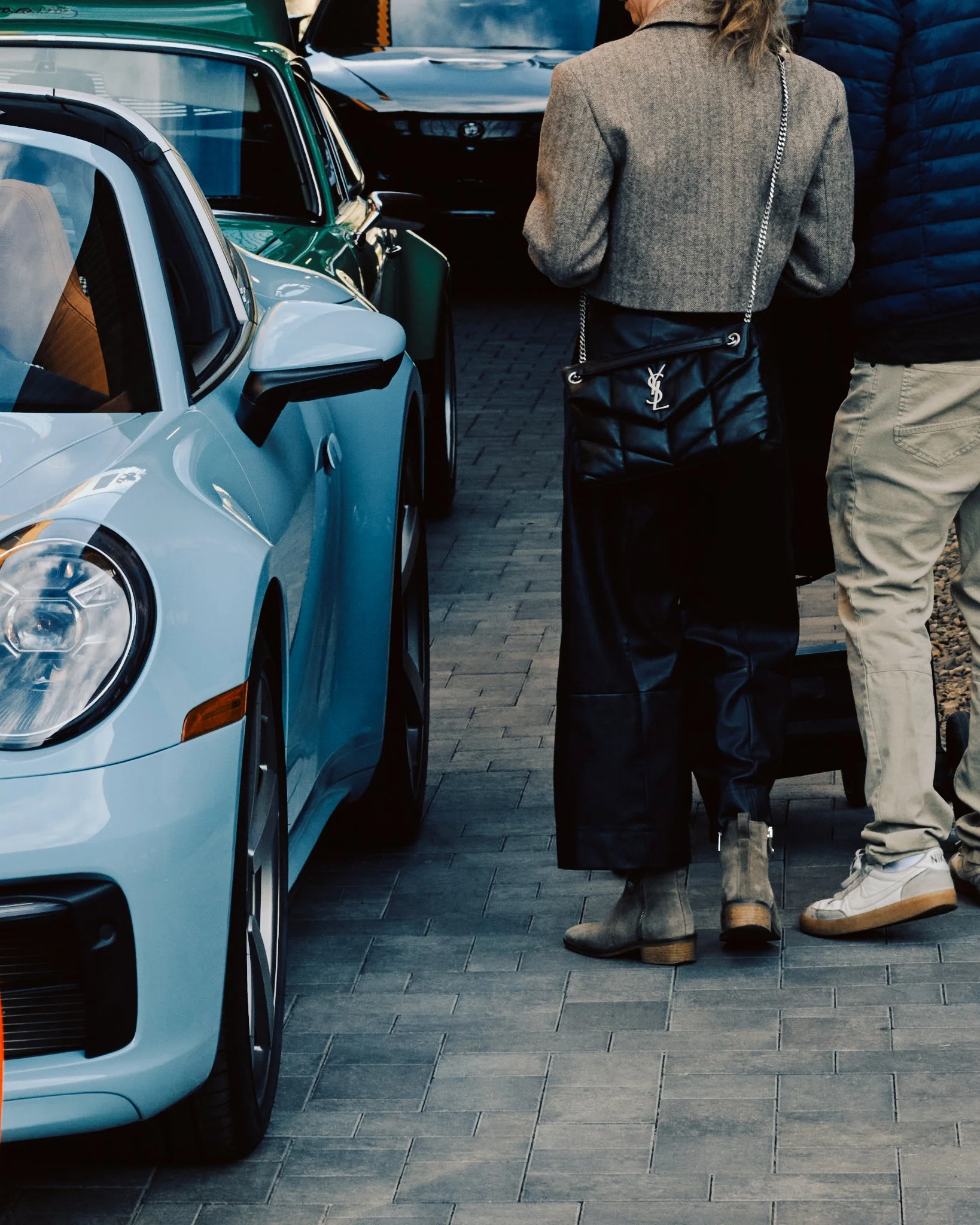 Two people standing on a brick sidewalk near parked cars, including a light blue sports car, engaged in conversation. One person wears black leather pants, a grey jacket, and carries a black Yves Saint Laurent purse. The other is dressed in a navy pu