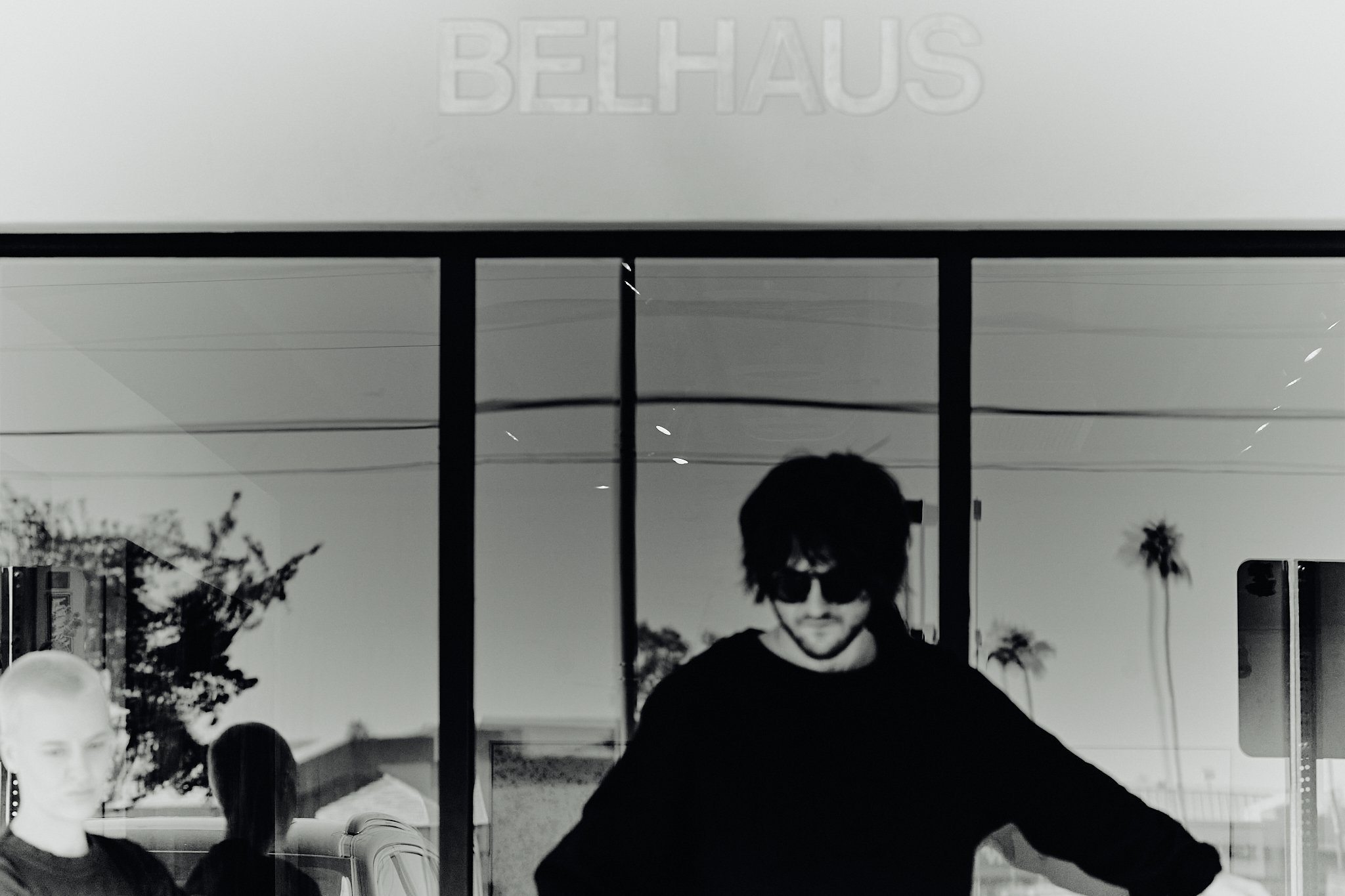 Reflection of a man with dark hair and sunglasses inside a building with the word "BELHAUS" above, and palm trees outside. A young woman with short blonde hair is also visible in the reflection.