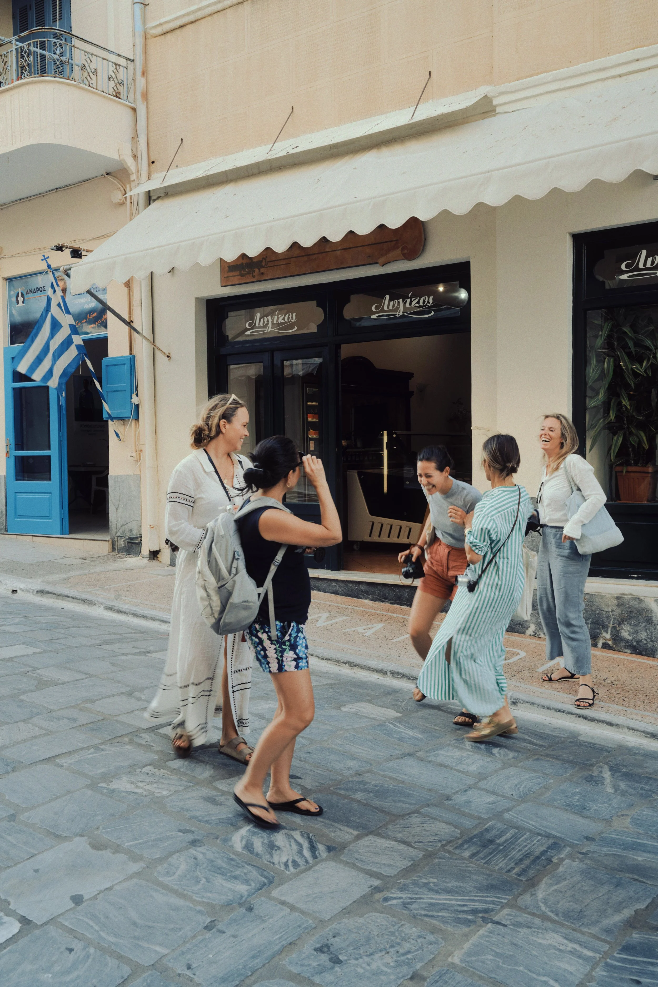 Group of five women laughing and enjoying a conversation on a cobblestone street outside a shop with a white awning and Greek flag