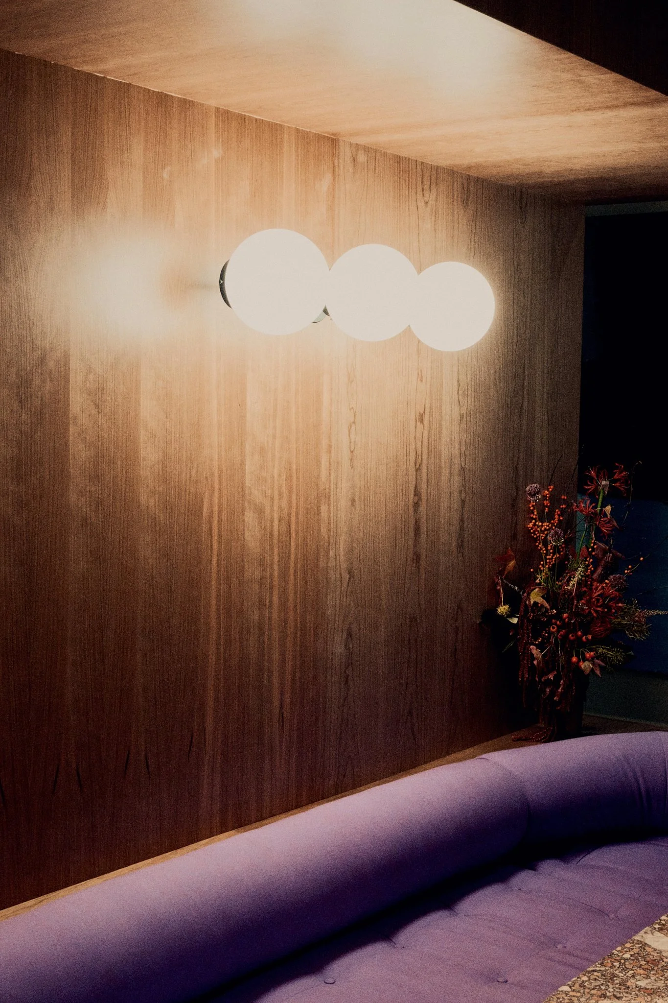 A modern room with a wooden wall, a contemporary white spherical wall sconce, a purple sofa, and a floral arrangement in a vase.