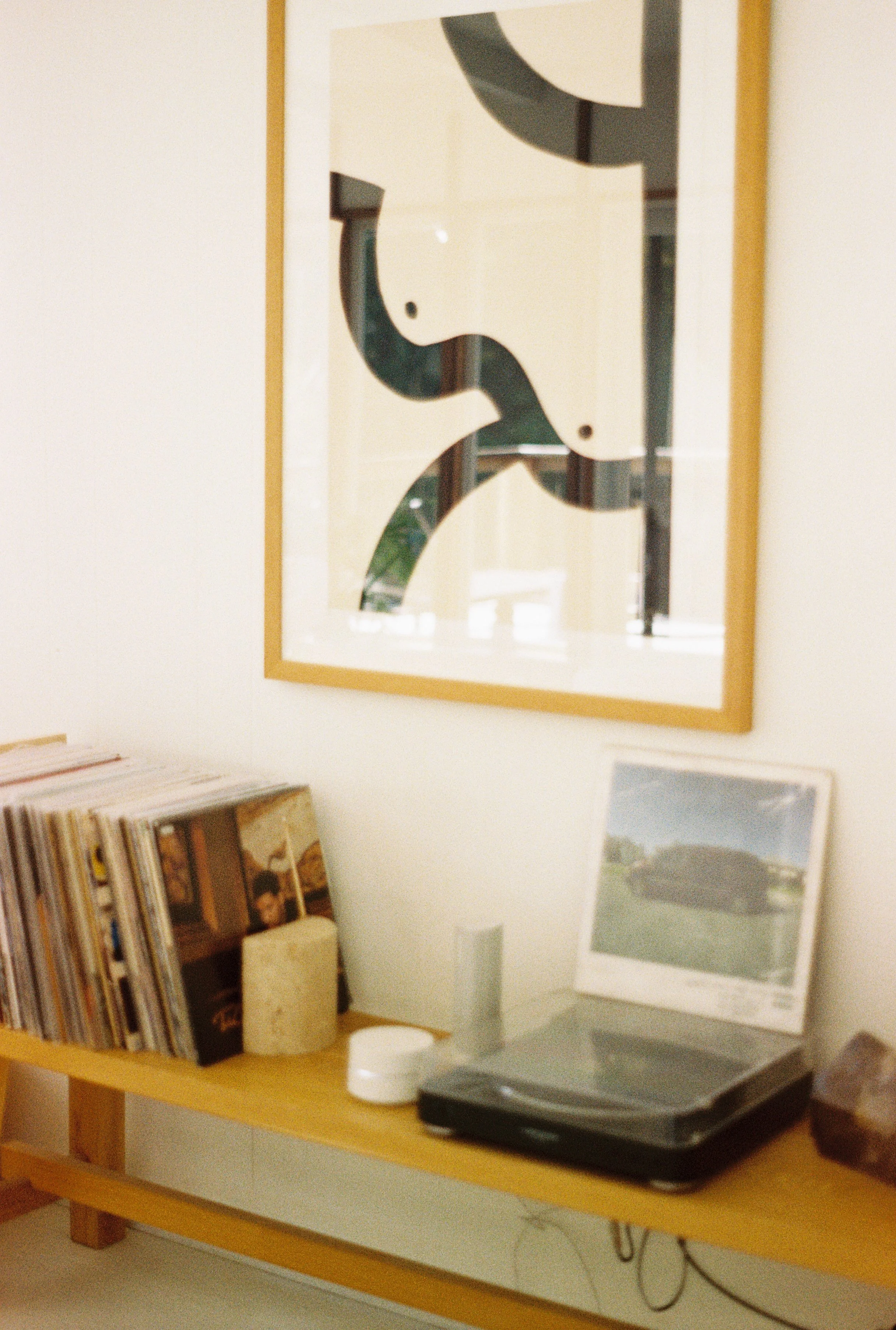 A wooden shelf holding vinyl records, a white speaker, a framed photo of a car, and a black electronic device next to a framed abstract art piece on the wall.