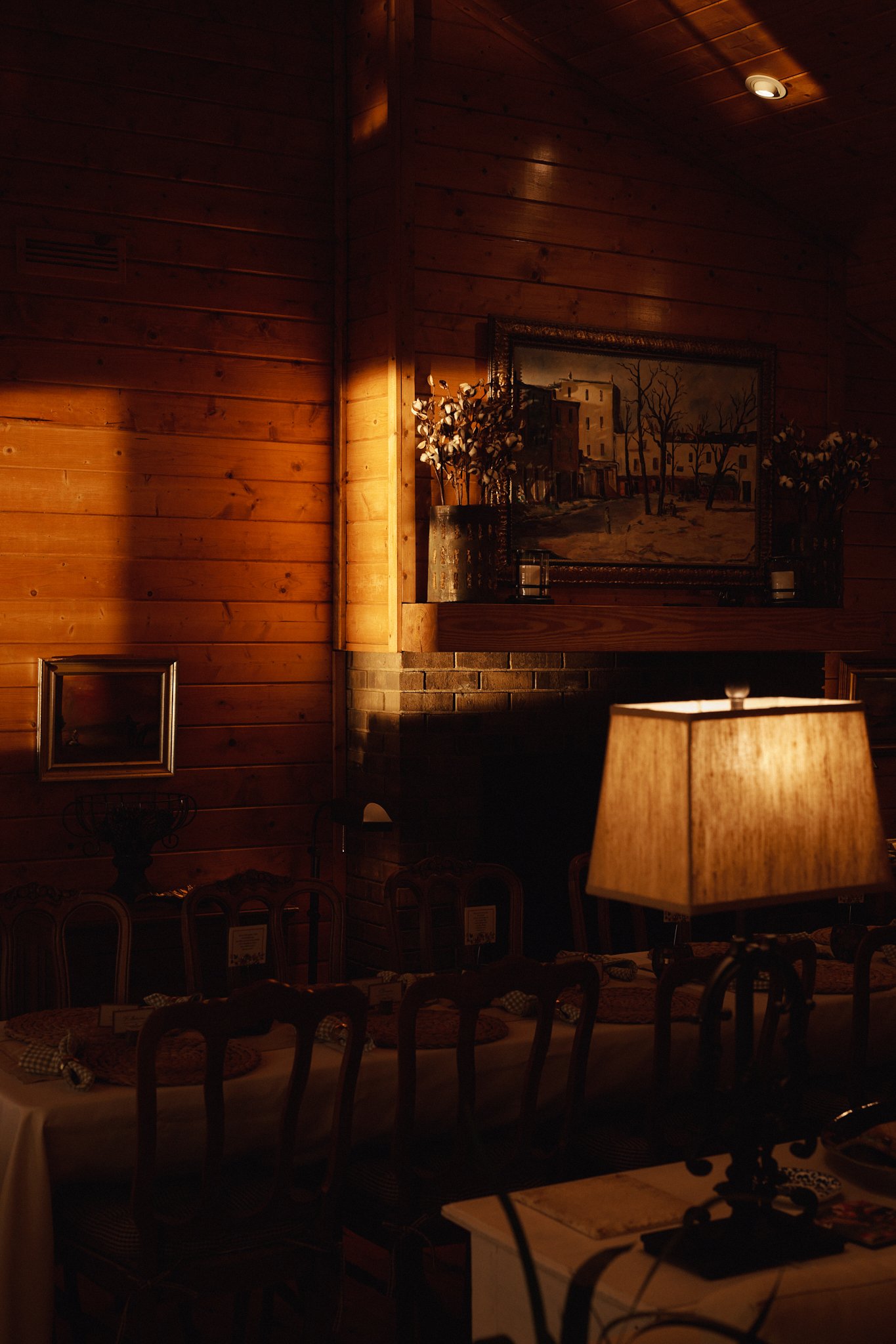 Interior of a cozy dining room with wooden walls, a table set with placemats, a table lamp, a fireplace, and framed artwork on the wall.