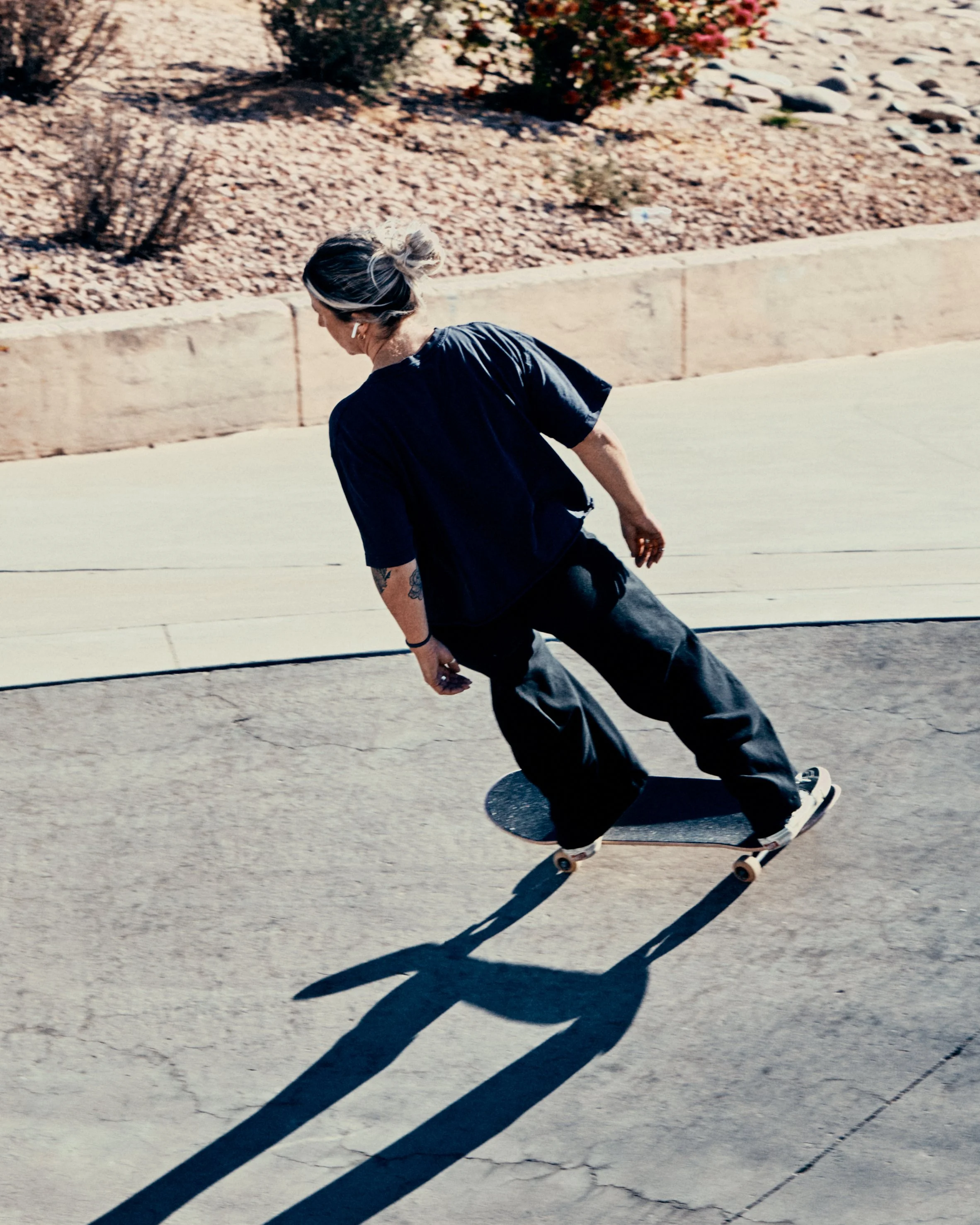 A person skateboarding on a paved surface with a shadow cast on the ground, a desert landscape with rocks and bushes in the background.