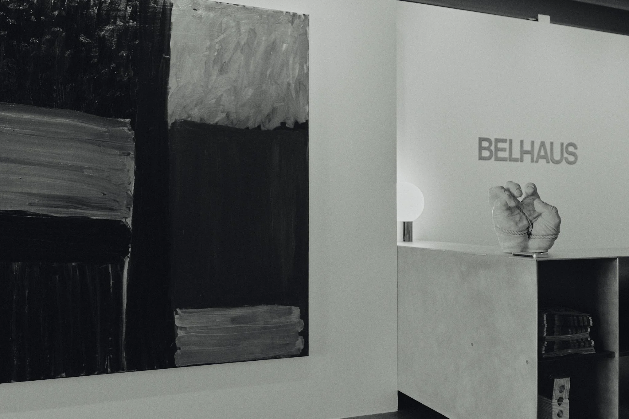 A white wall with a black and gray abstract painting on the left and a white shelf with a sculpture and the word 'BELLHAUS' on the right in a modern interior setting.