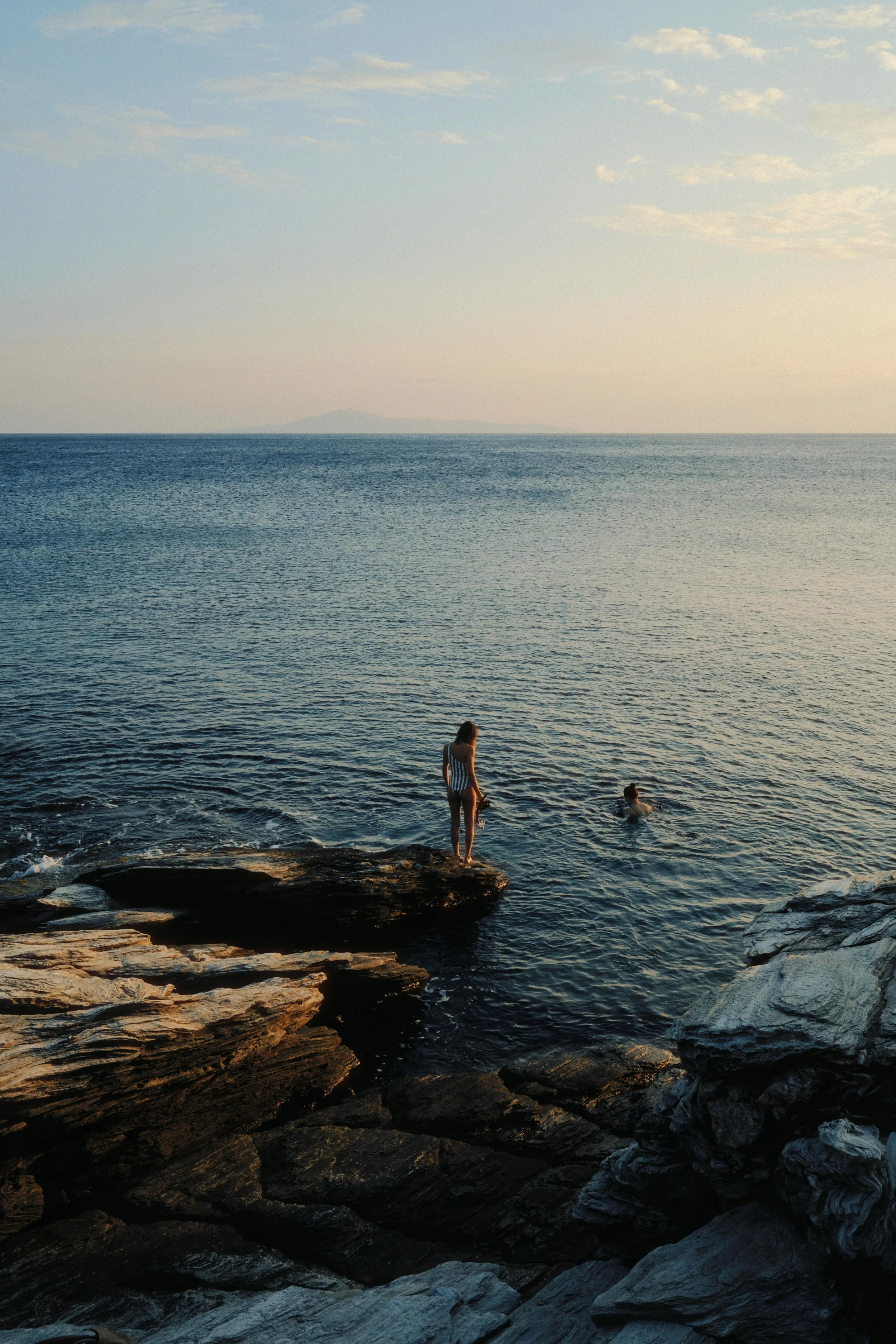 A woman standing on a rock ledge at the edge of the ocean while another person swims nearby during sunset, with calm water and a distant island on the horizon.