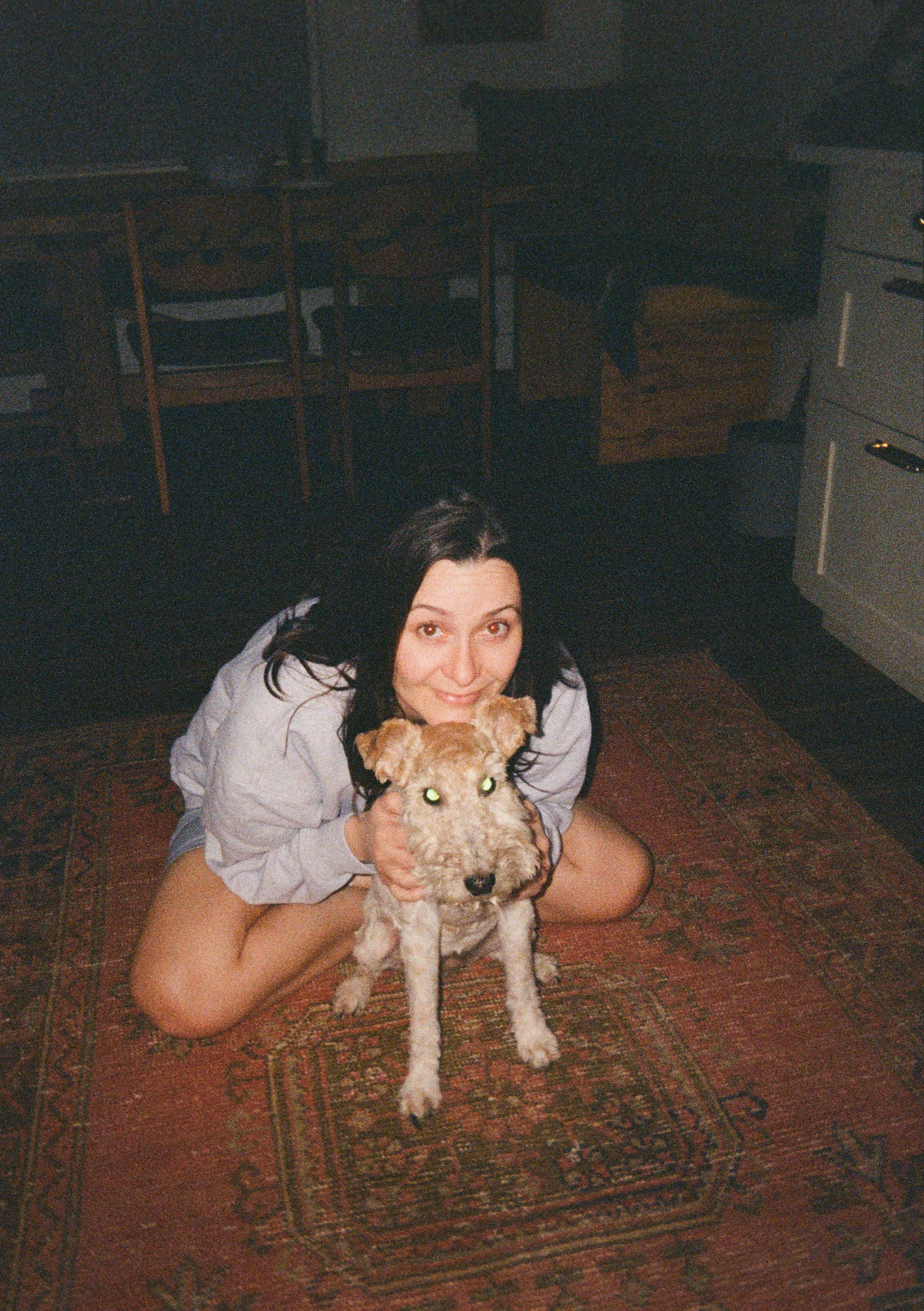 A woman with dark hair, smiling, squatting on a patterned rug indoors, holding an alert dog with a scruffy coat in front of her.