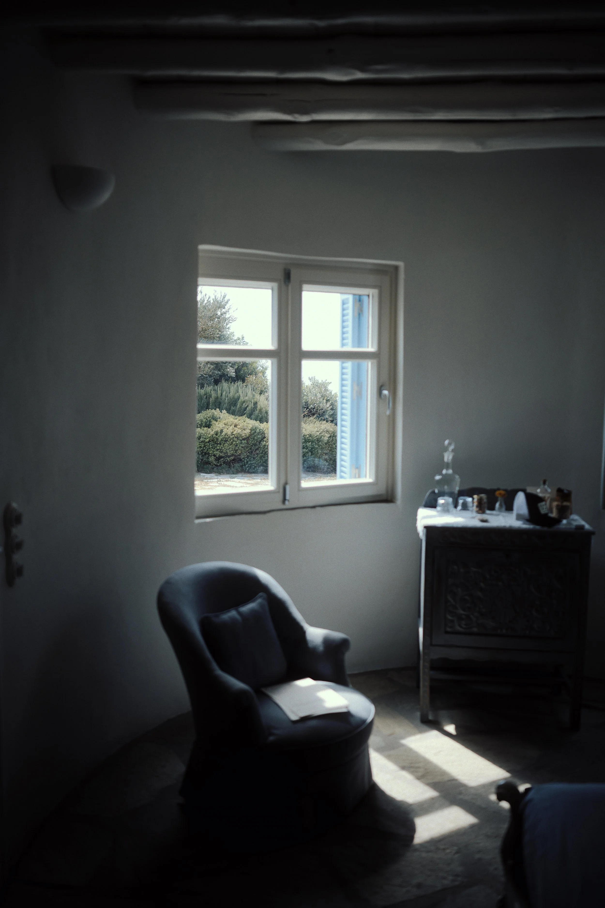 A dimly lit room with a window showing an outdoor view of greenery and a blue building. The room contains a dark armchair with a pillow and a paper on it, and a decorative black cabinet with glassware and bottles on top.
