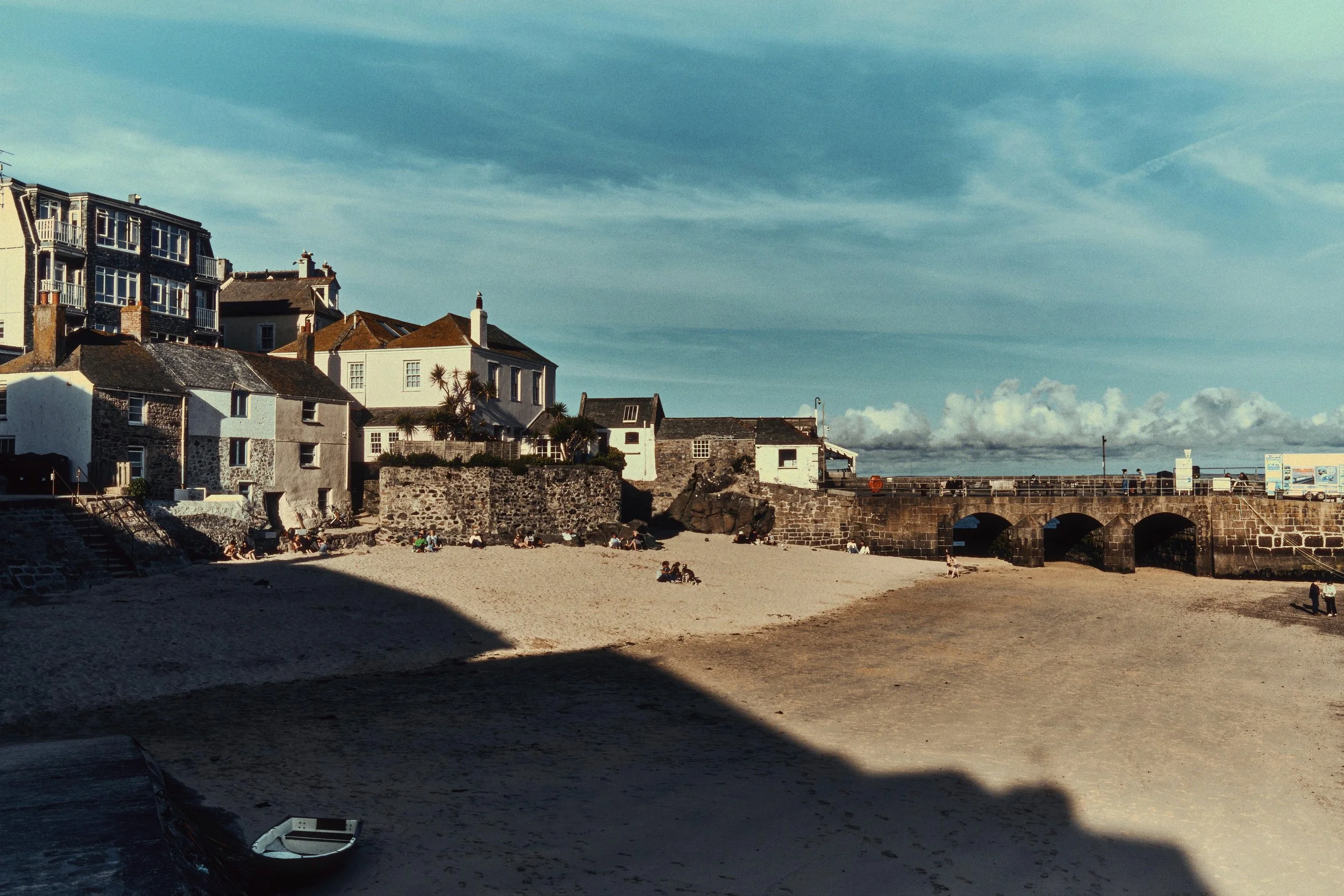 Beach with sand, a stone bridge, and coastal buildings under a partly cloudy sky.