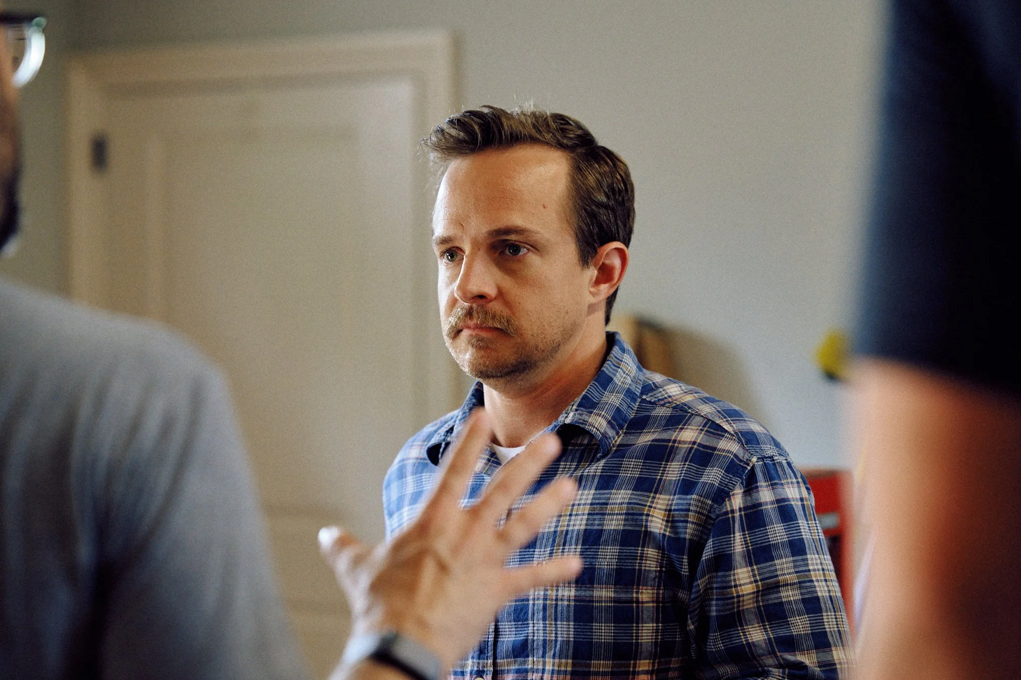 A man with brown hair and a mustache wearing a blue plaid shirt looks serious while talking to a person with a tattooed arm, in an indoor setting.
