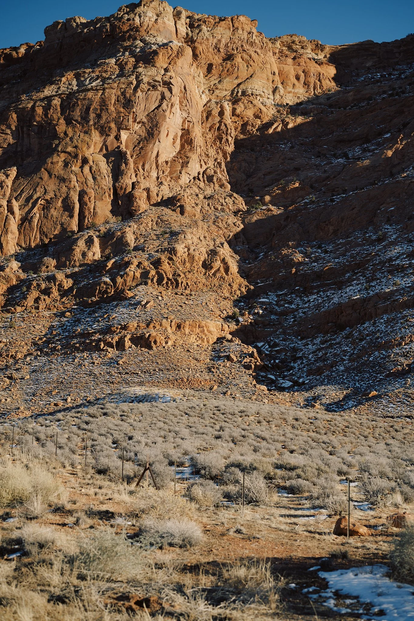 A rocky desert landscape with sparse bushes and patches of snow, featuring tall reddish-brown cliffs under a clear blue sky.