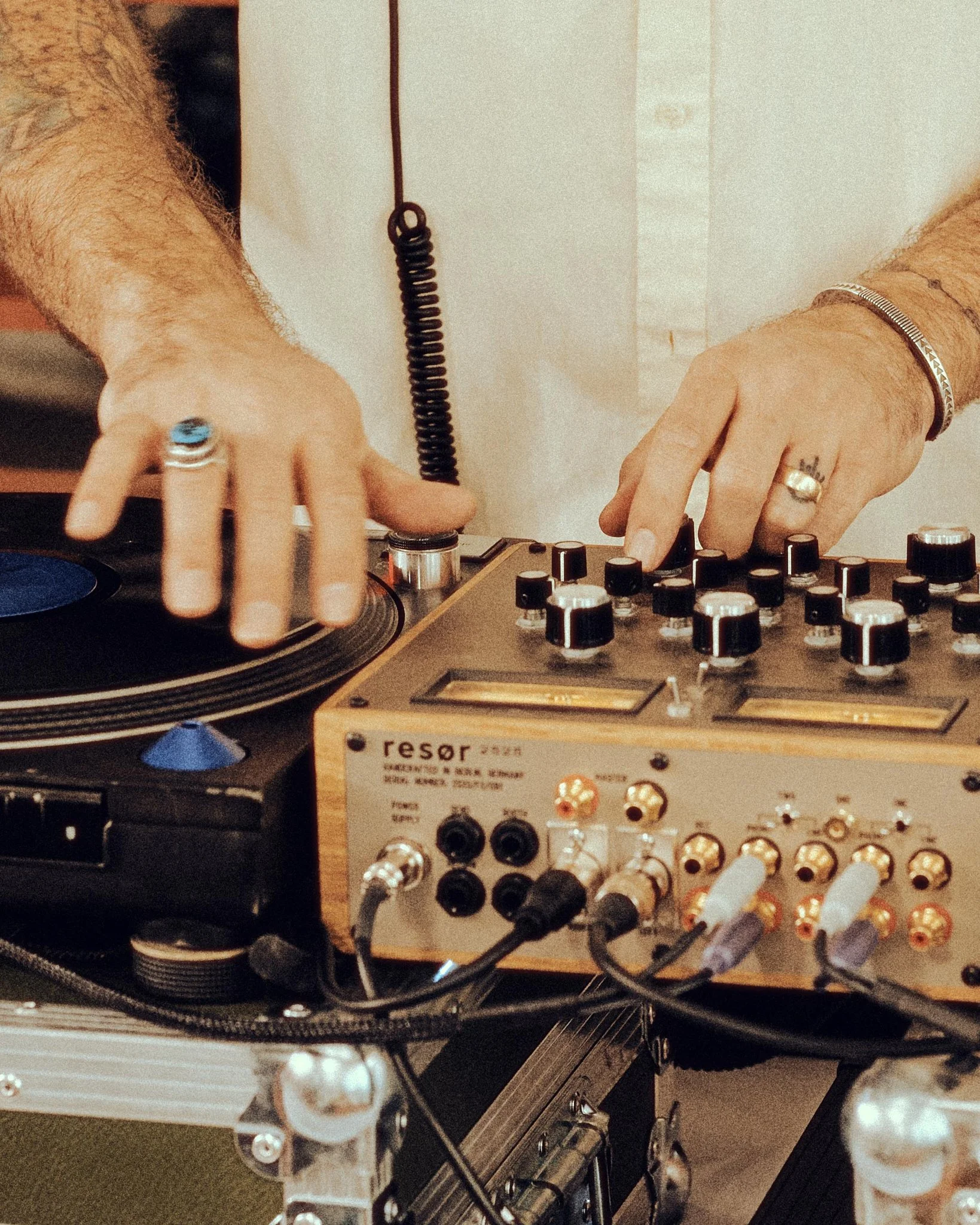 Close-up of a DJ's hands, one with a large ring and the other with a bracelet, operating a turntable and a resistor audio interface surrounded by various cables and equipment.