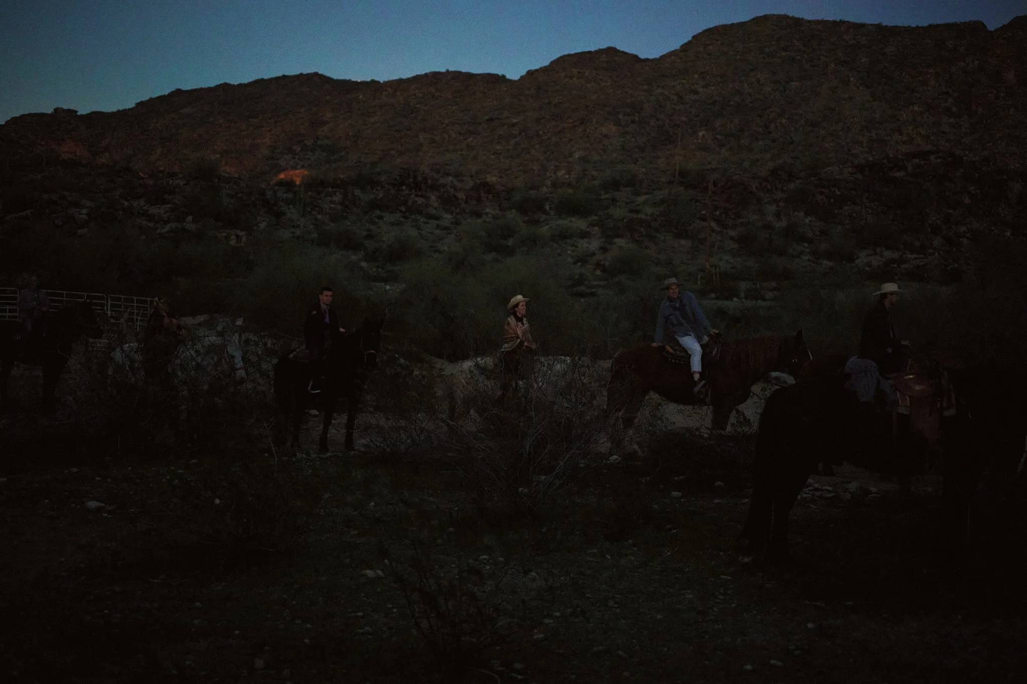 A group of people riding horses in a desert landscape at dusk, with hills in the background.
