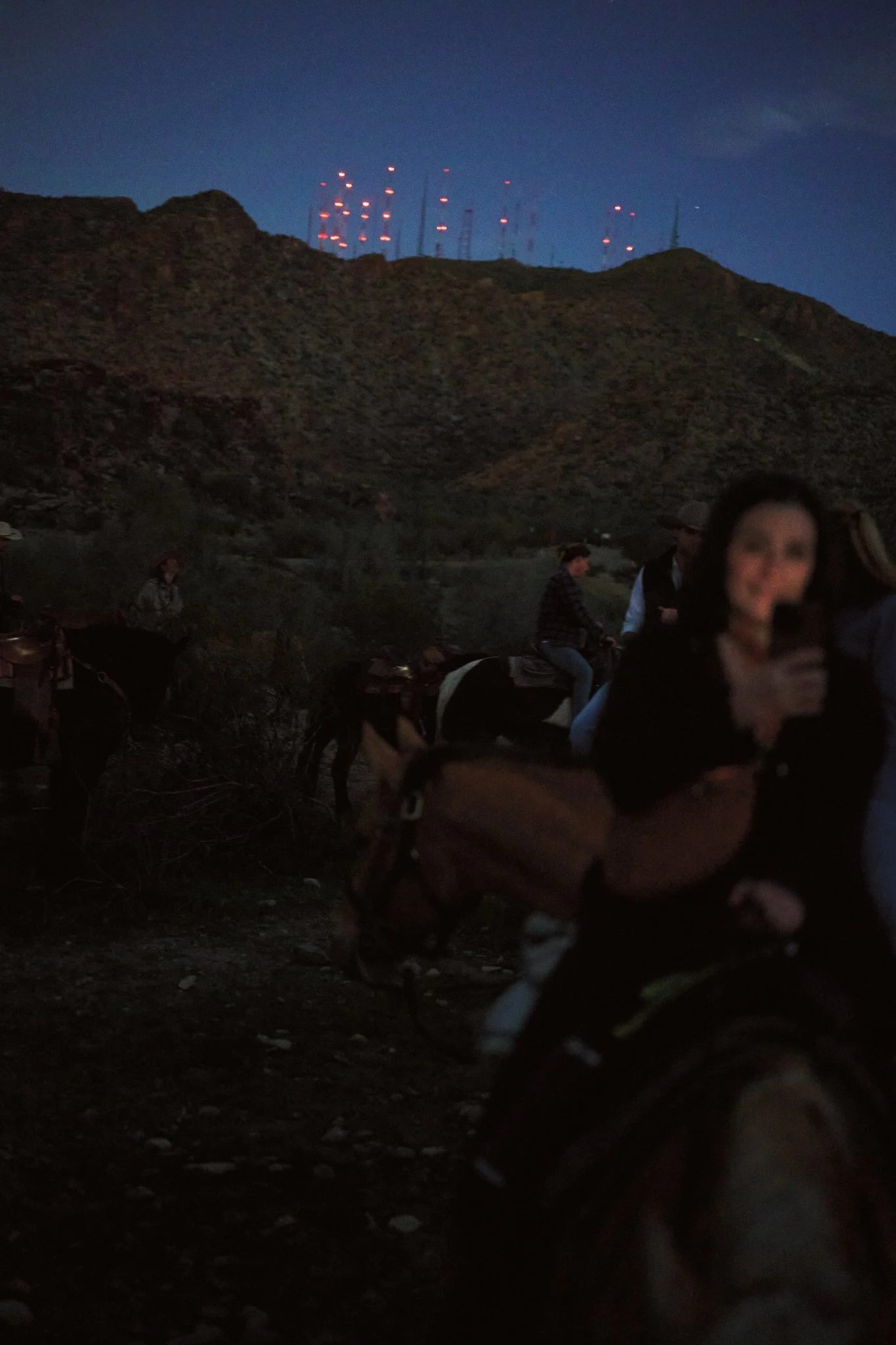 People riding horses outdoors at night in a mountainous area, with radio towers on a hilltop in the background.