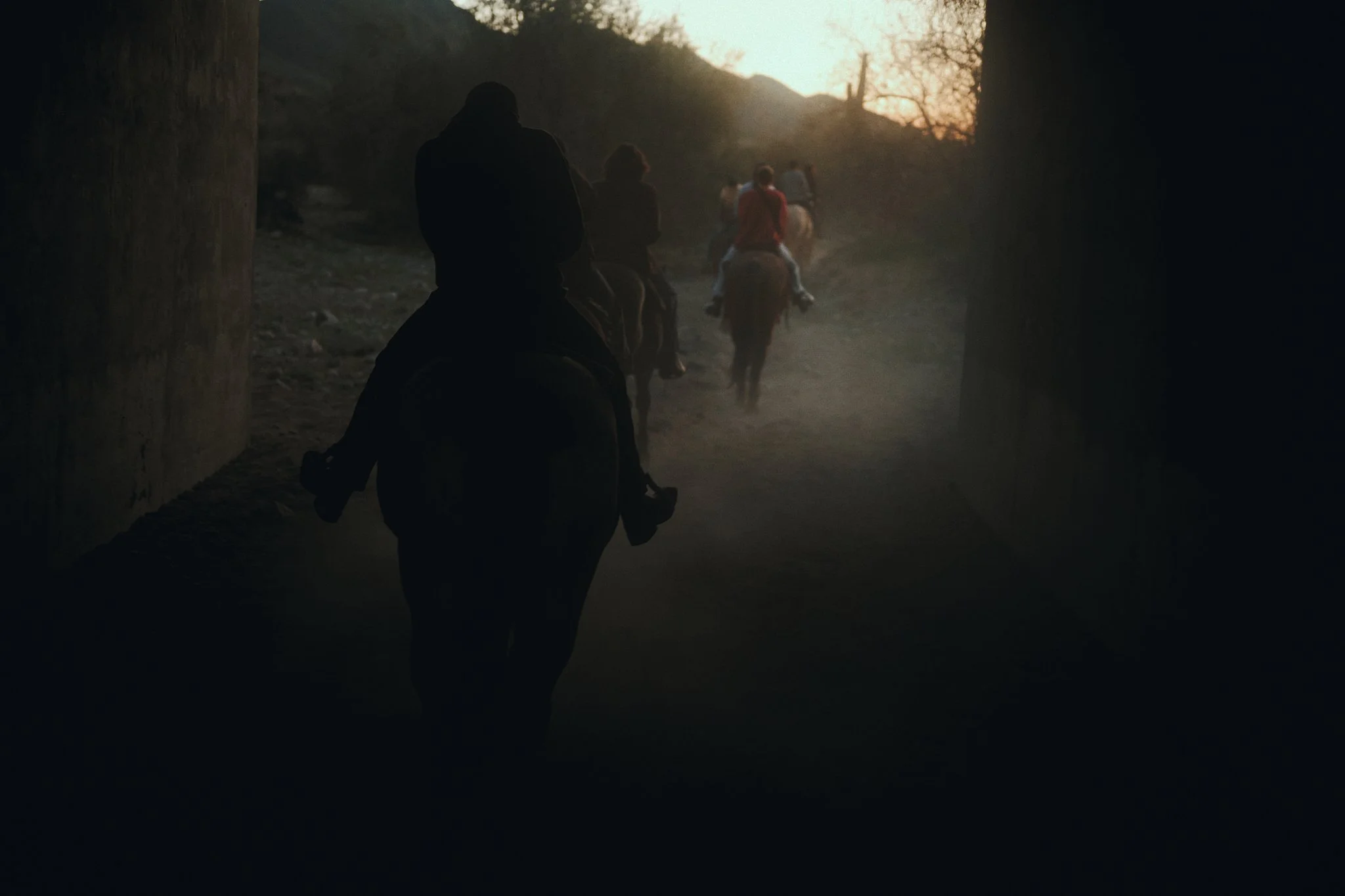 Silhouettes of people riding horses under a bridge at sunset, with mountains in the background.