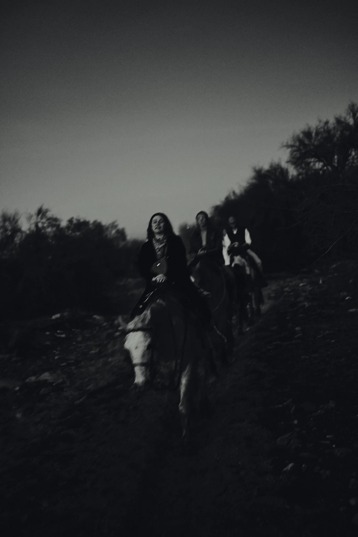 Three people riding horses at dusk or night, in a dark outdoor setting with trees in the background.
