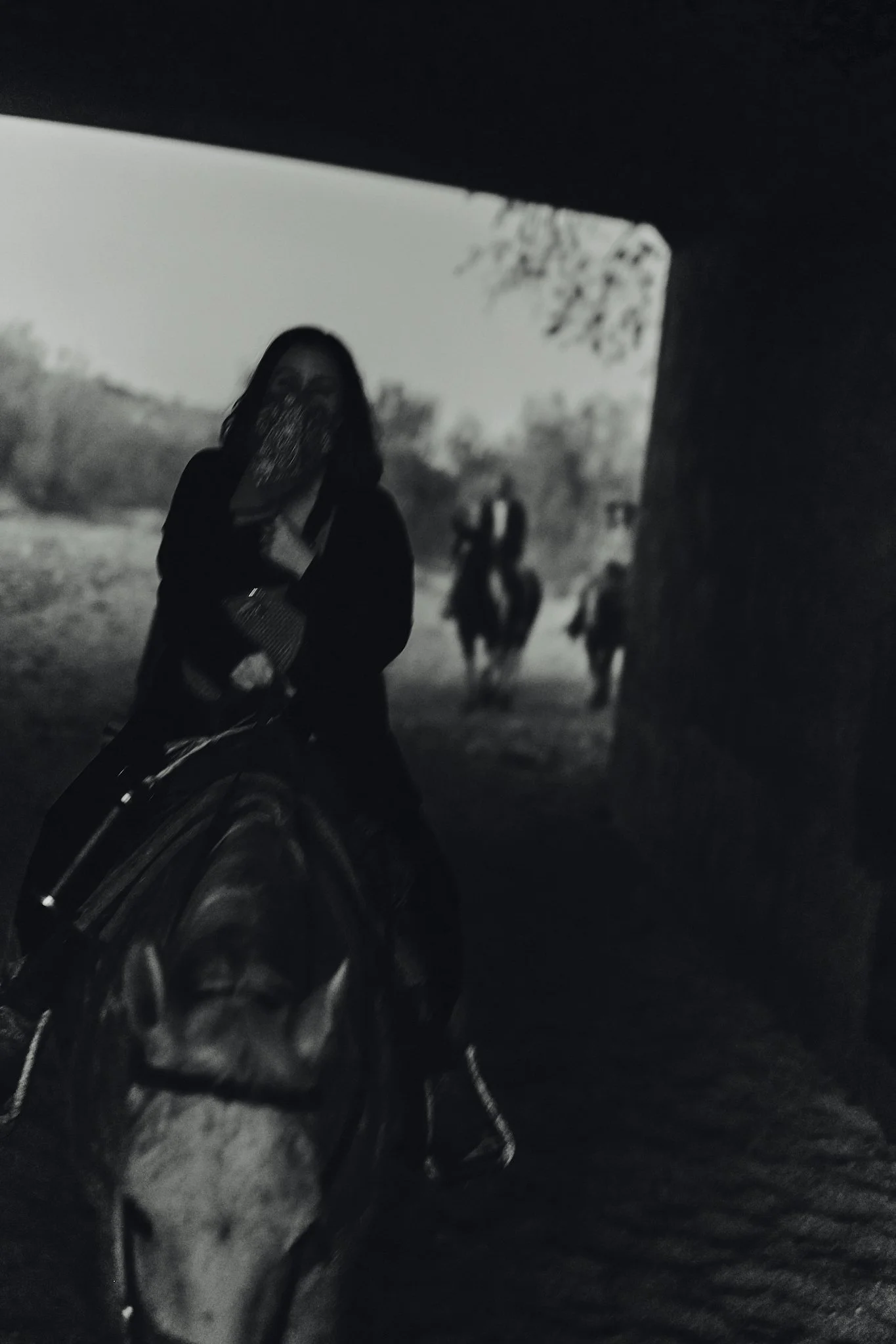 A person with long hair wearing a face mask on horseback, with two other horses and riders in the background, outdoors near a tree.