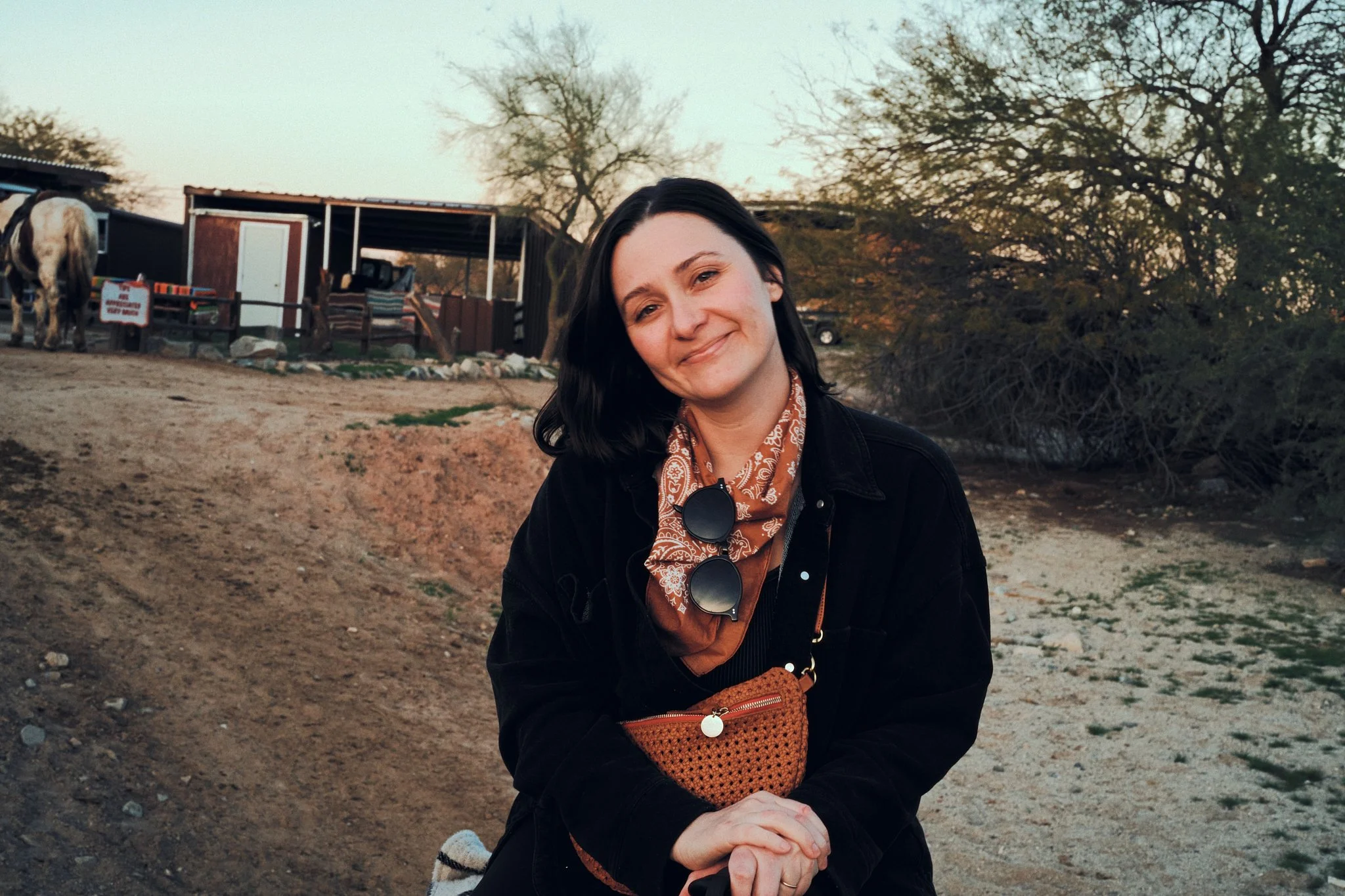 A woman with black hair smiling, wearing a black jacket and a brown bandana, sitting on a dirt path outdoors with trees and a small rustic building in the background.