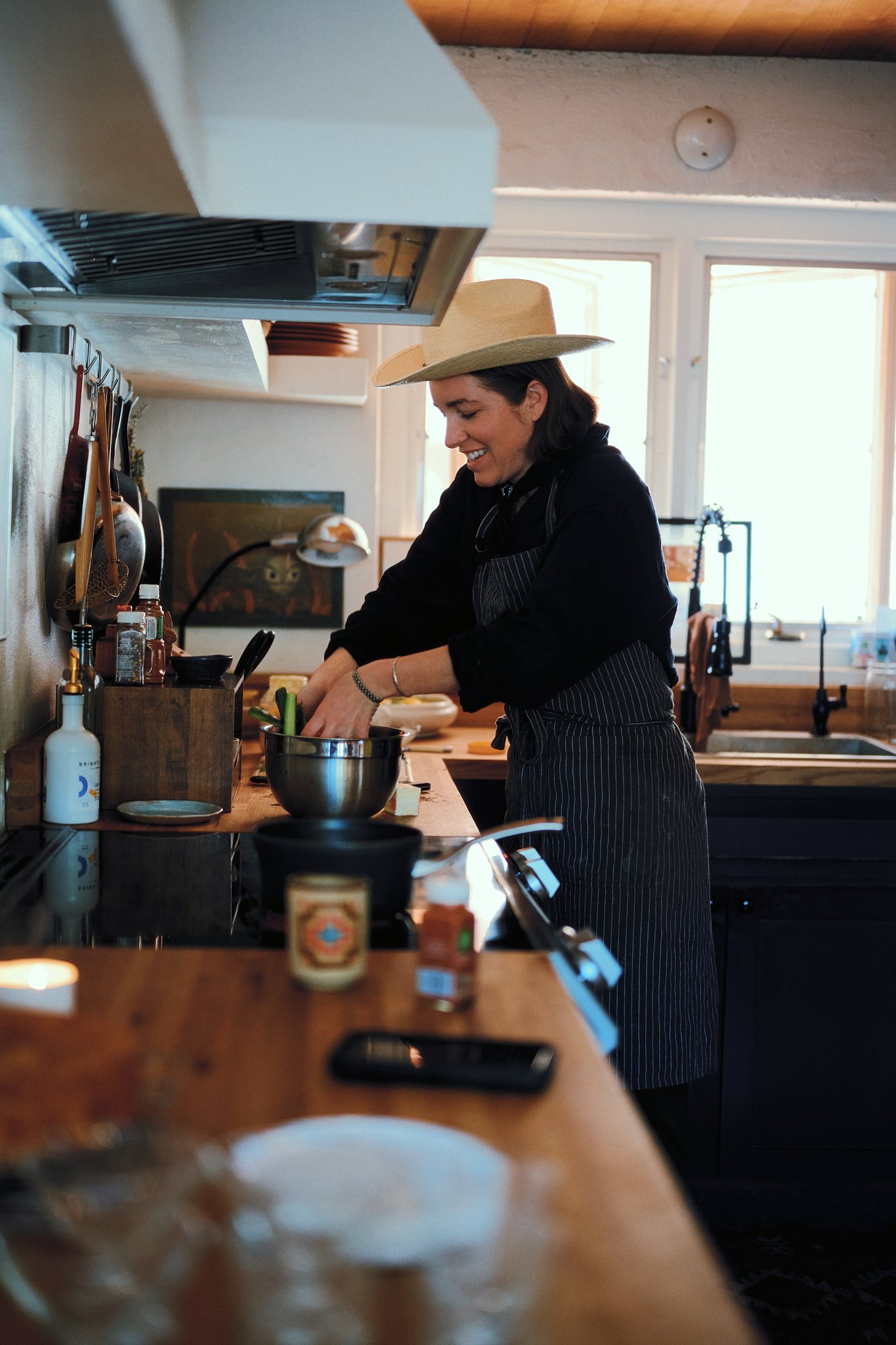A woman wearing a straw hat and a black apron is cooking in a cozy kitchen with natural light, standing at a wooden counter.