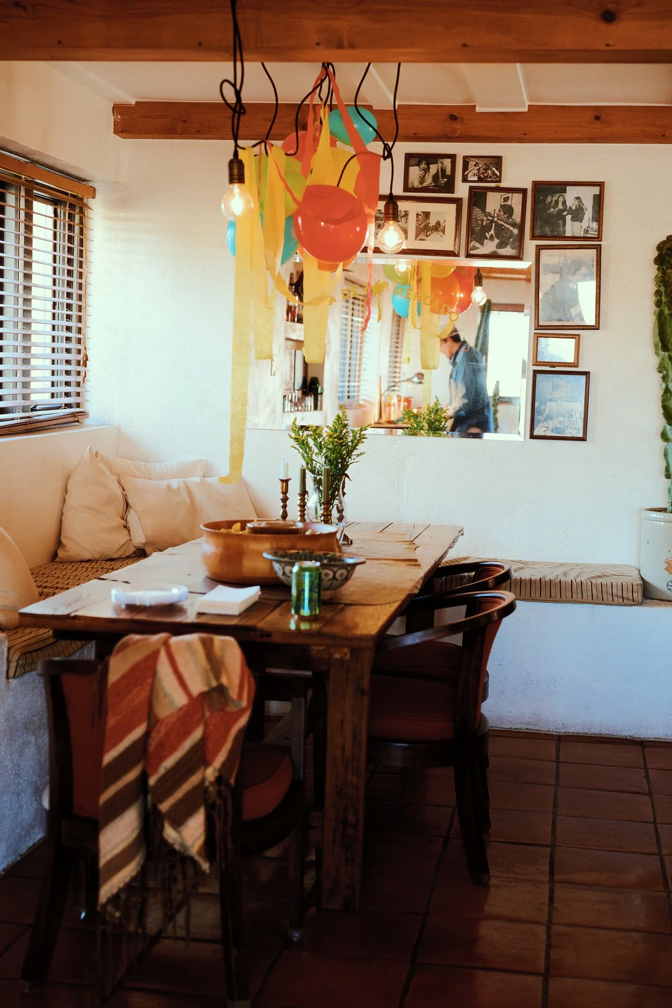 Decorated dining area with balloons and streamers hanging from the ceiling, a wooden table with chairs, a bench with cushions, framed photos on the wall, and a person visible in the kitchen reflected in a mirror.