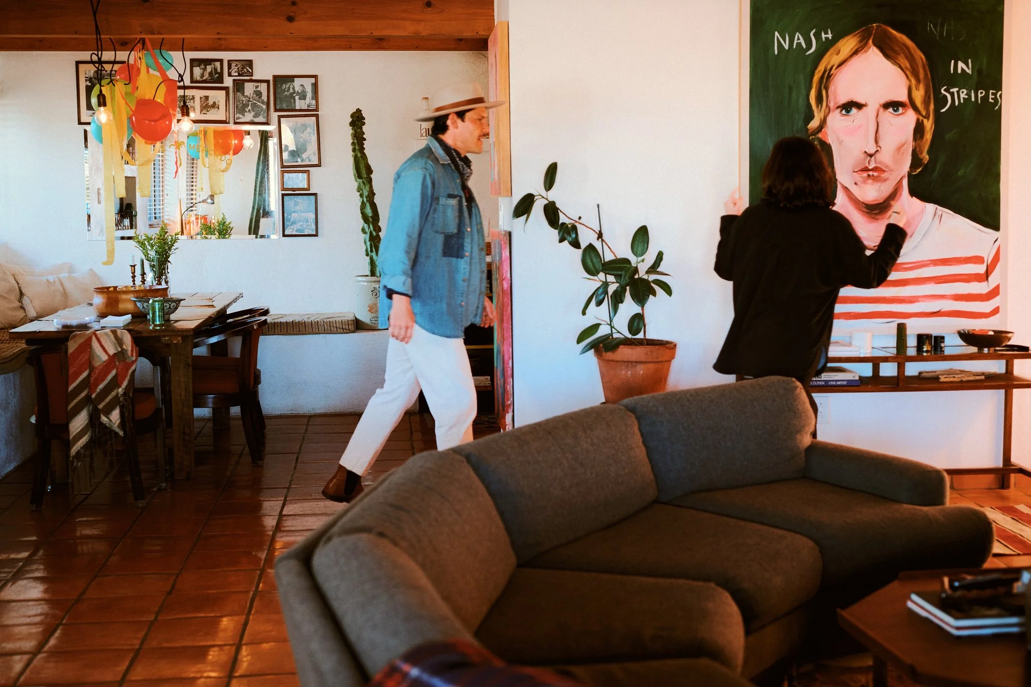 A woman is painting a portrait of a man on a large canvas in a cozy living room, while a man wearing a straw hat and denim jacket walks past her. The room has a brown couch, a dining table with chairs, and decorated walls with photographs and artwork